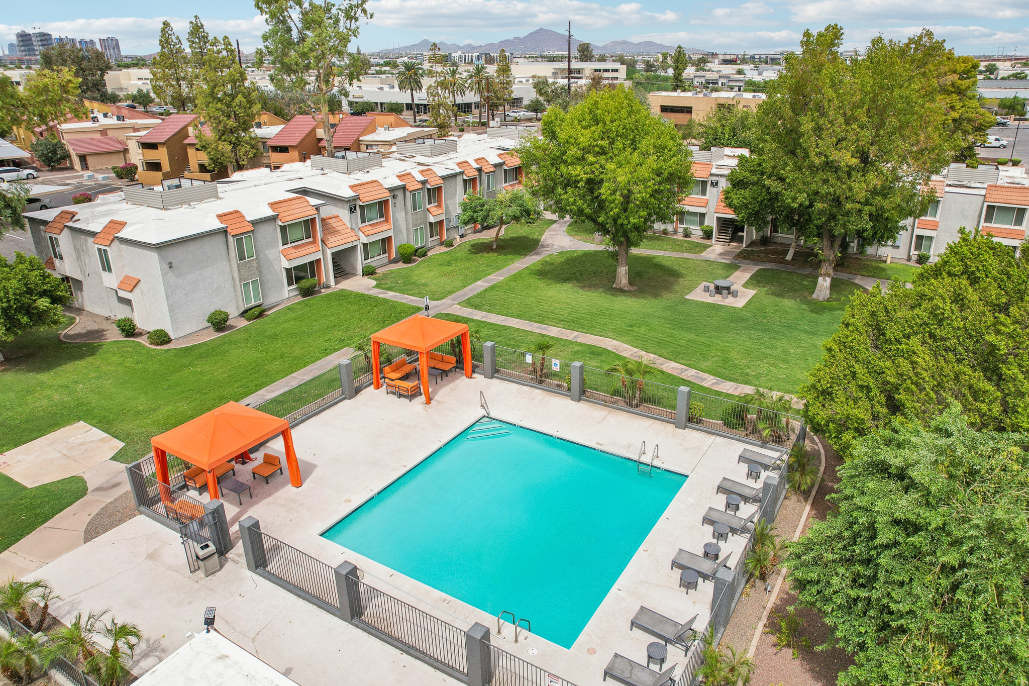 Aerial view of a swimming pool surrounded by palm trees and lounge chairs. Two orange cabanas are beside the pool, which is situated within a landscaped area. In the background, two-story residential buildings and green trees are visible, with a cloudy sky overhead.