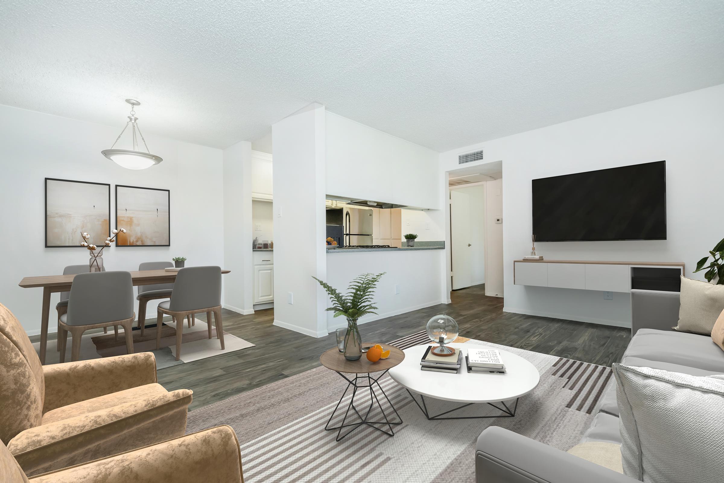 A modern living room featuring a comfortable seating area with two chairs, a gray sofa, and a coffee table. The space includes a dining area with a table and chairs, and a kitchen visible in the background. Light walls and flooring create a bright and open atmosphere, complemented by decorative plants and wall art.