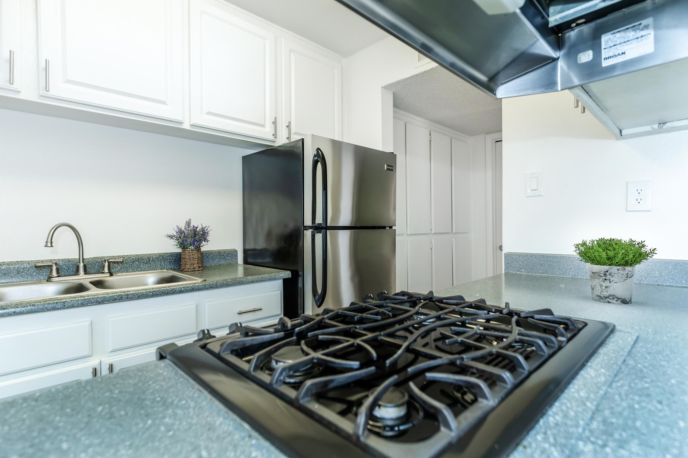 A modern kitchen with white cabinets, a stainless steel refrigerator, and a gas stove. The countertops are gray with a subtle texture, and there are small decorative plants on the counter. The kitchen is well-lit and features a clean, organized layout.