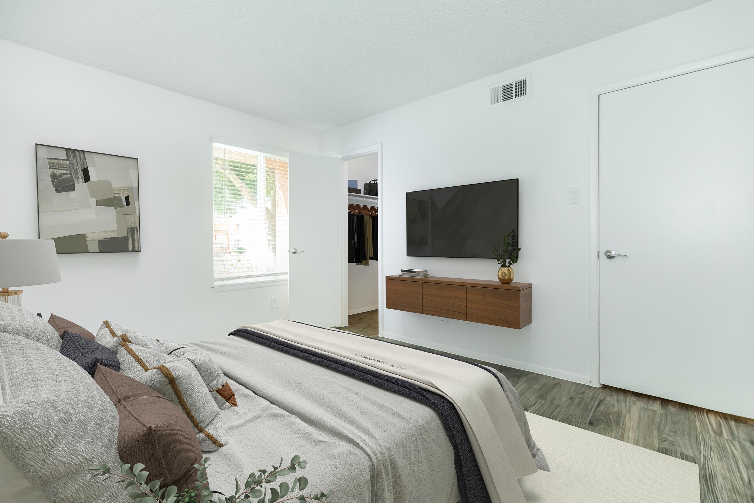 A modern bedroom featuring a neatly made bed with decorative pillows, a minimalist artwork on the wall, a mounted flat-screen TV, and a wooden media console. A light-colored rug covers the floor, and a daylight window illuminates the space. A closet door is visible in the background.