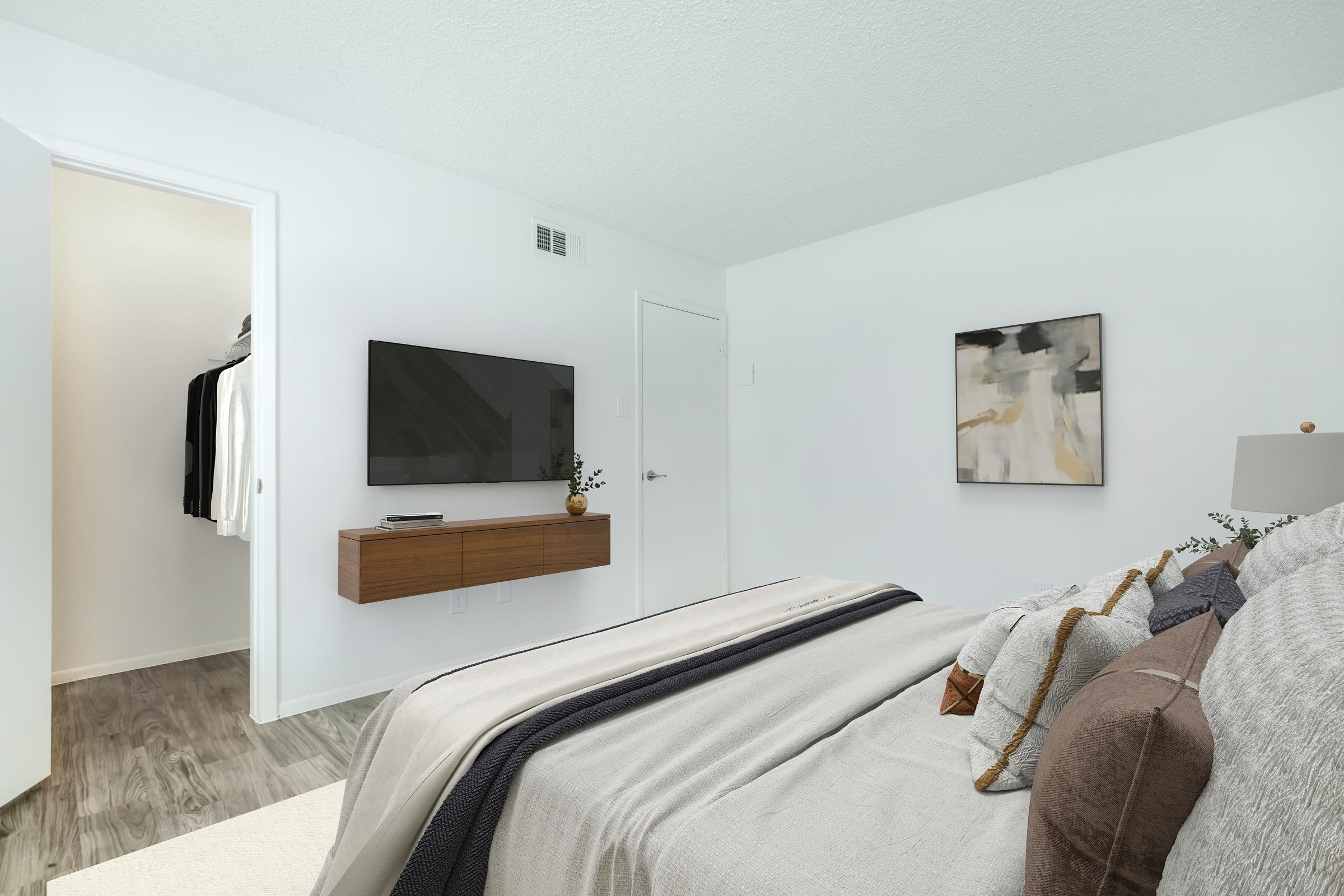 A modern bedroom featuring a cozy bed with decorative pillows, a mounted flat-screen TV on the wall, and a wooden dresser underneath. A closed closet door is visible on the left, and a piece of abstract artwork hangs above the dresser. The room has light-colored walls and a warm, inviting atmosphere.