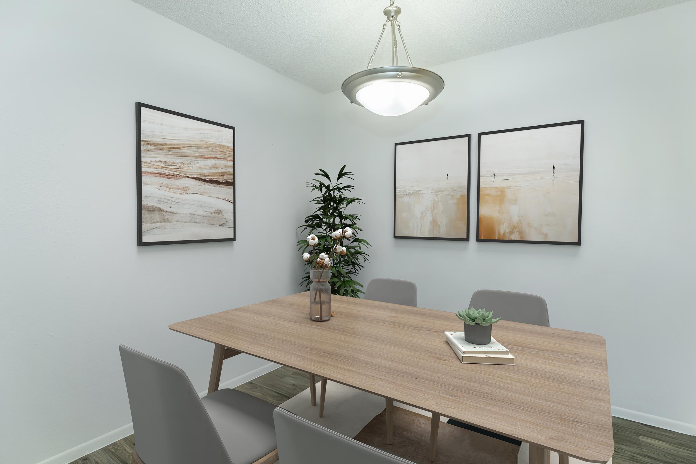 A modern dining area featuring a wooden table surrounded by gray chairs, a vase with flowers, and a small succulent plant. Two abstract framed artworks are hung on the white walls, and a green plant adds a touch of nature to the space. A stylish light fixture hangs from the ceiling.