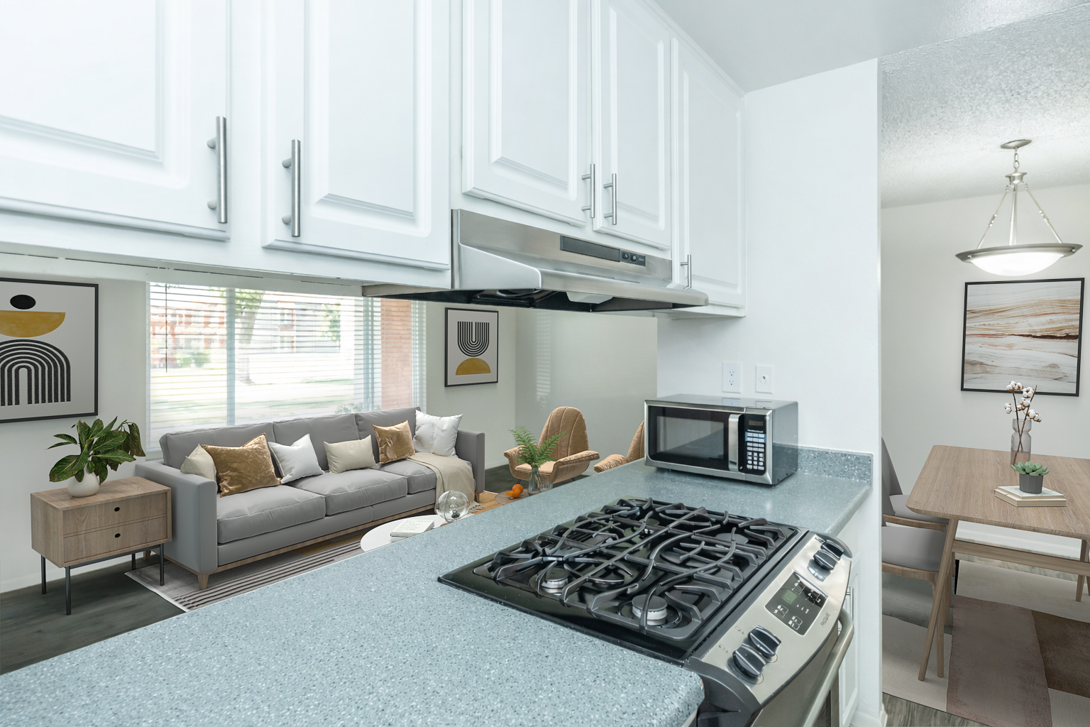 A modern kitchen featuring white cabinets, a gas stove, and a countertop with a microwave. In the background, a living area is visible, showcasing a gray sofa with colorful cushions, a wooden coffee table, and a dining area with a wooden table and chairs, illuminated by natural light from a window.