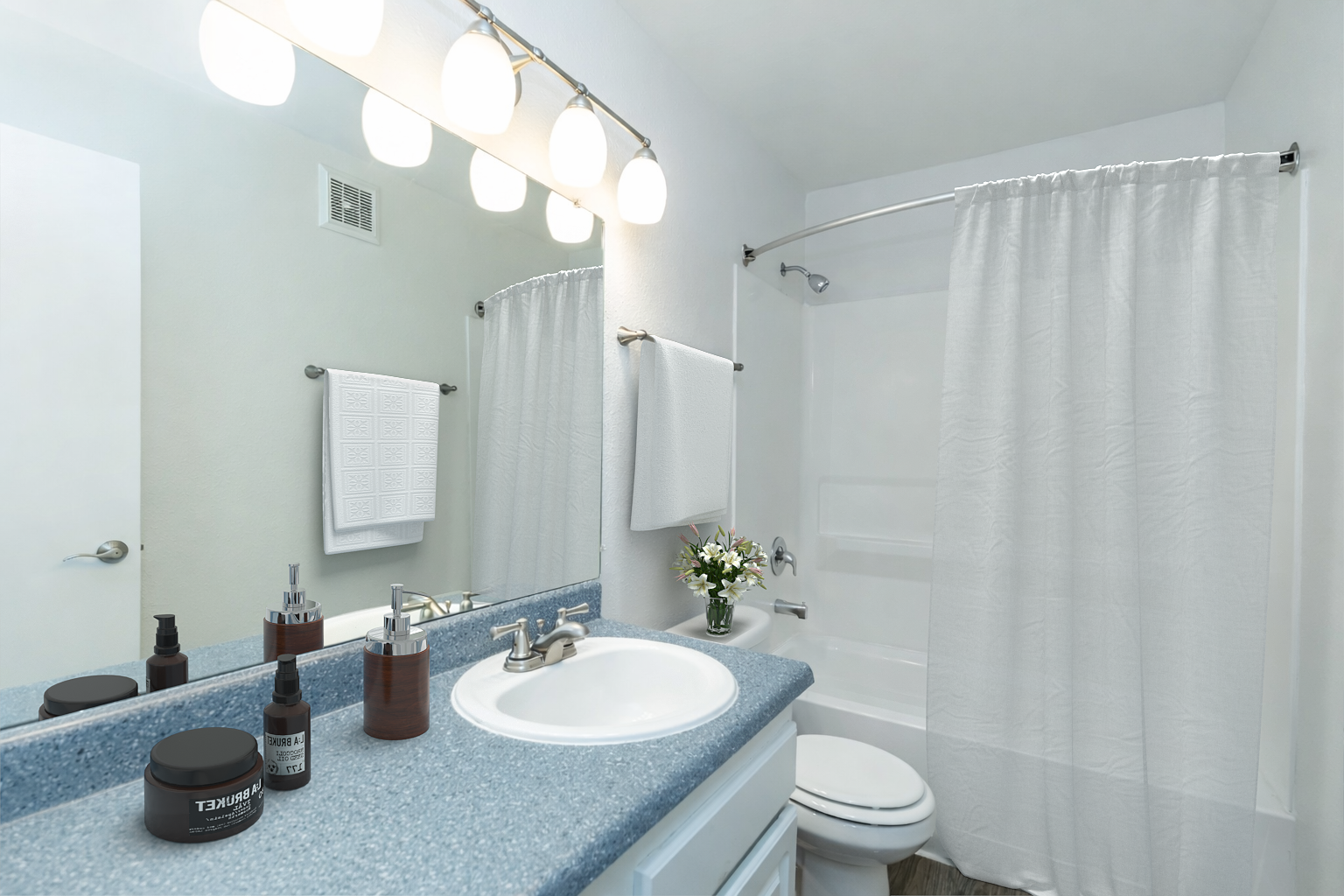 A clean, modern bathroom featuring a sink with toiletries, a mirror above the sink, a shower curtain, and fresh flowers. The walls are light-colored, and a white towel is hanging on a rod. Soft lighting from above illuminates the space. The overall ambiance is tidy and inviting.