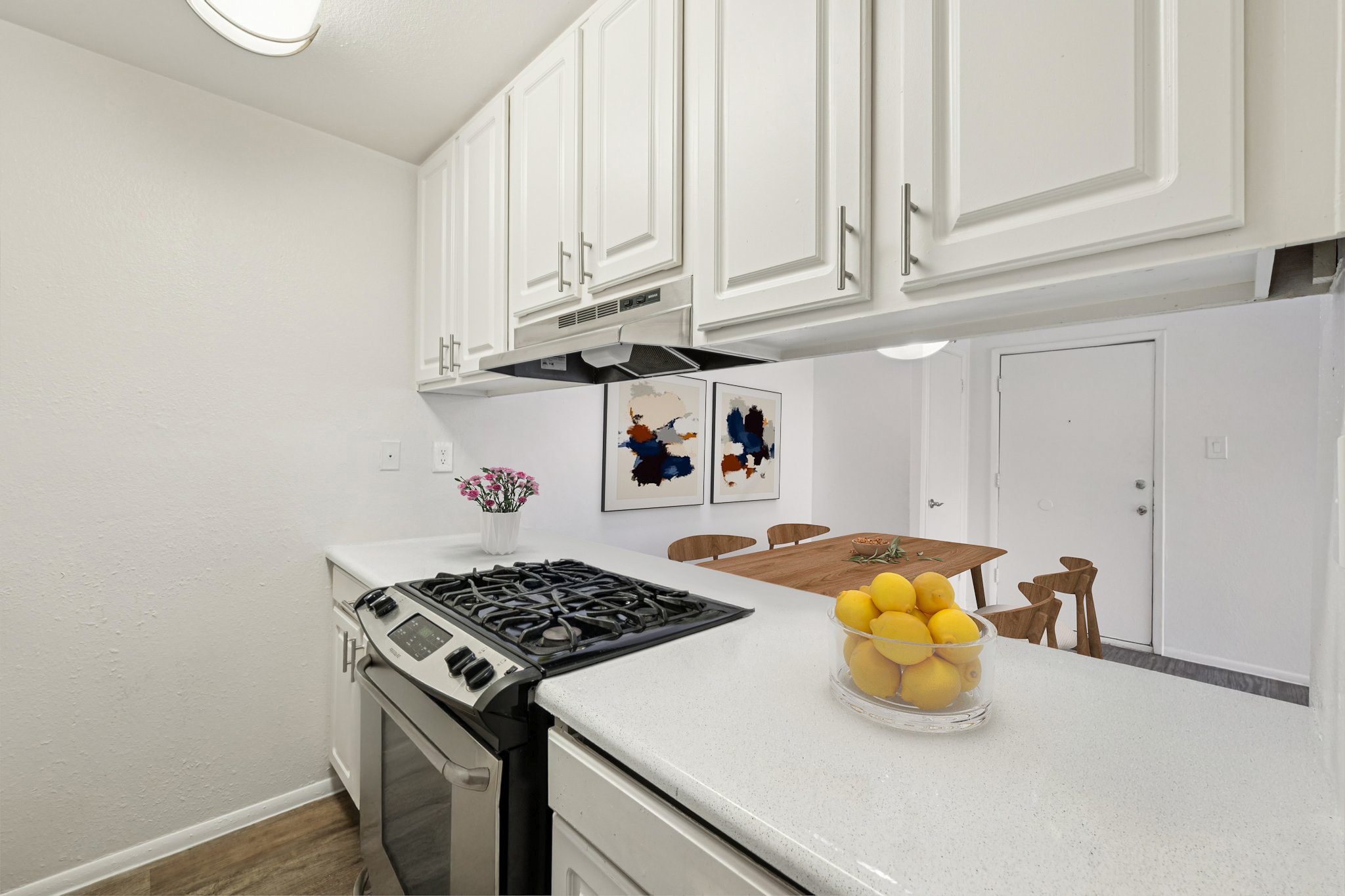 A modern kitchen featuring white cabinetry, a gas stove, and a countertop with a bowl of lemons. In the background, there is a dining table with wooden chairs and two framed abstract artworks on the wall. Flowers in a vase add a touch of color, and a doorway is visible in the background.
