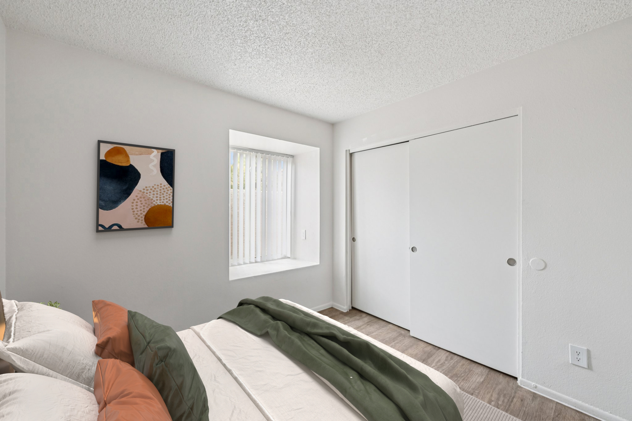 A cozy bedroom featuring a bed with beige and green bedding, a piece of abstract wall art, a window with vertical blinds, and a closed closet door. The walls are painted in neutral tones, and the floor has light-colored laminate, creating a bright and inviting atmosphere.