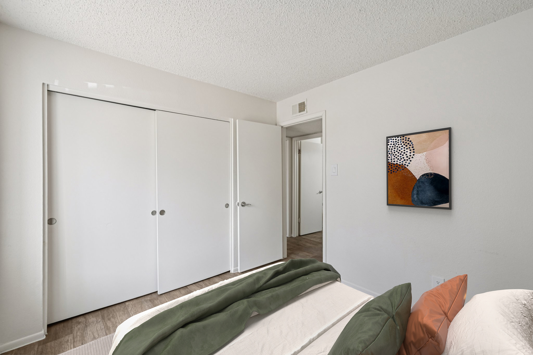 A well-lit bedroom featuring a bed with green bedding, light-colored walls, and a minimalist design. On the wall, there's an abstract framed artwork. Two closed white closet doors are visible, and a doorway leads to another room. The overall ambiance is clean and modern.