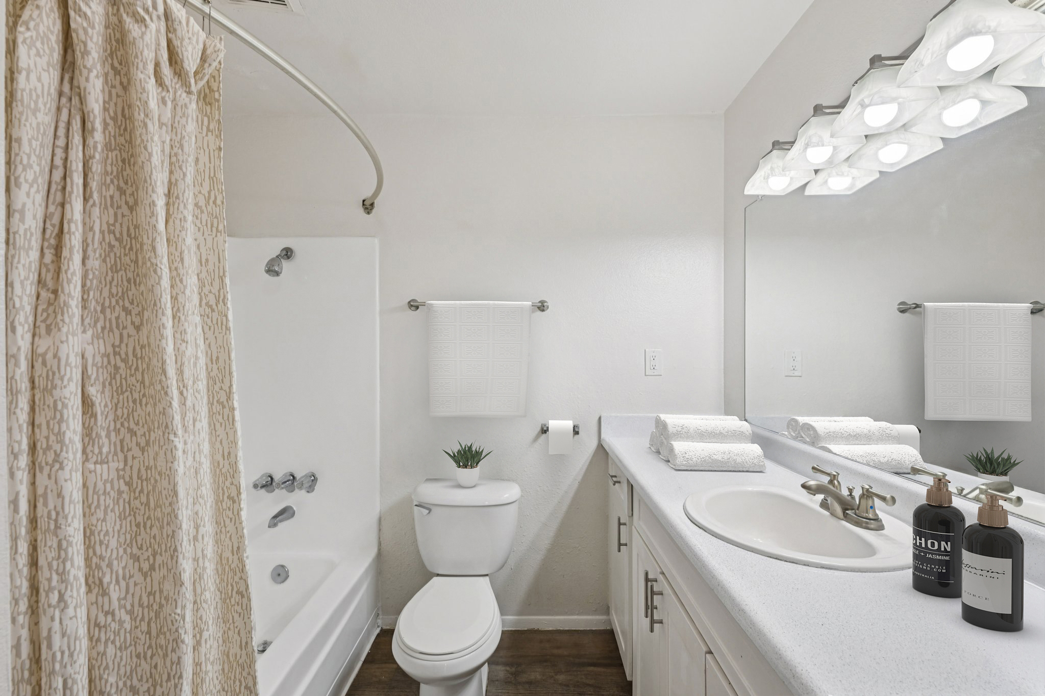 A clean, modern bathroom featuring a white bathtub with a shower curtain, a white vanity with a sink, and a large mirror with bright lighting. Two towels are neatly arranged on the countertop alongside decorative bottles and a small plant, creating a fresh and inviting atmosphere.