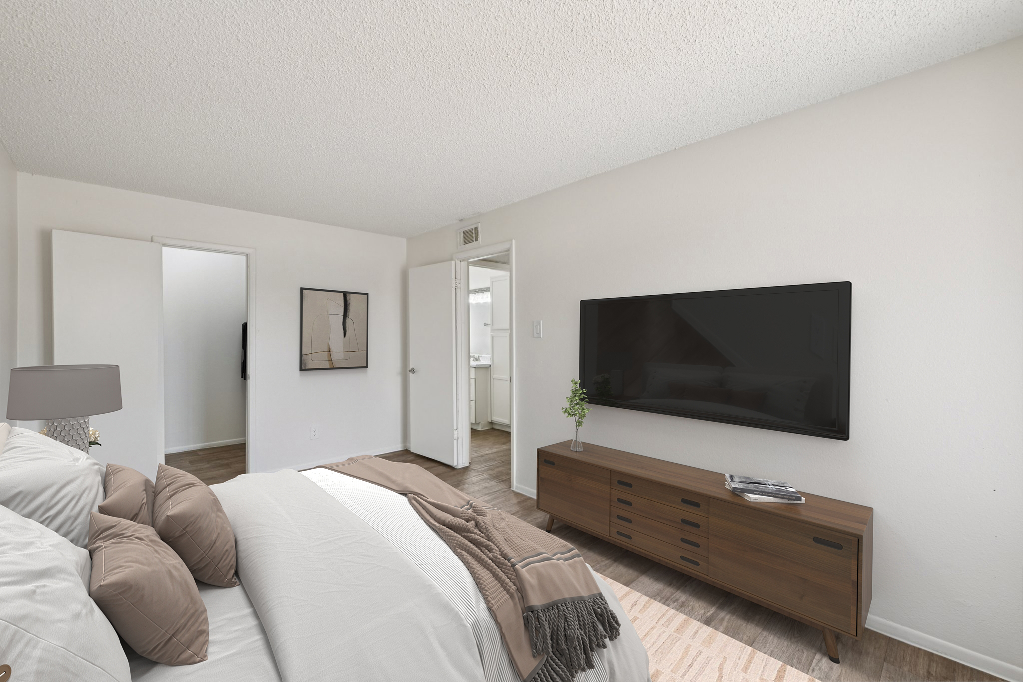 A cozy bedroom featuring a queen-sized bed with neutral-colored bedding, a modern wooden dresser with a flat-screen TV mounted on the wall, and soft lighting from a bedside lamp. The room has light-colored walls and flooring, with a doorway leading to a bathroom visible in the background.