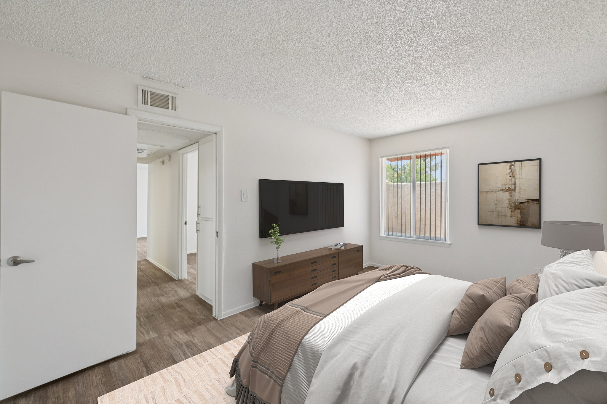 A cozy bedroom featuring a bed with light bedding and decorative pillows, a wooden dresser with a flat-screen TV mounted on the wall, and a small plant. Natural light streams in through a window with sheer curtains, and a doorway leads to another room. The walls are painted in neutral tones.
