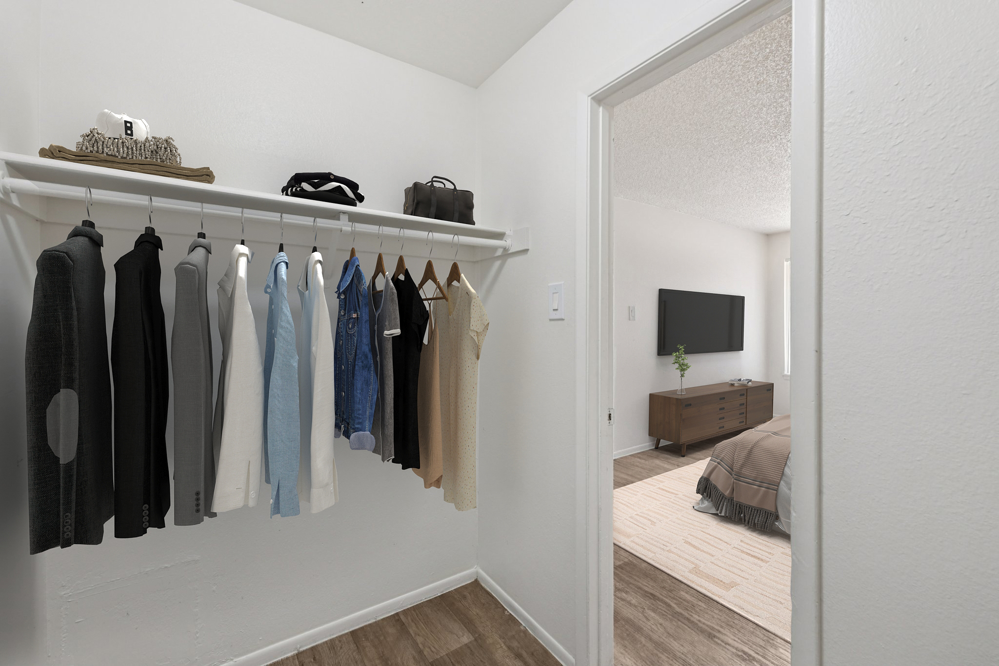 A well-organized wardrobe with a variety of shirts and jackets hanging on a white rack. In the background, a glimpse of a stylish bedroom is visible, featuring a TV and a modern dresser. The walls are painted white, and the flooring is a light brown wood.