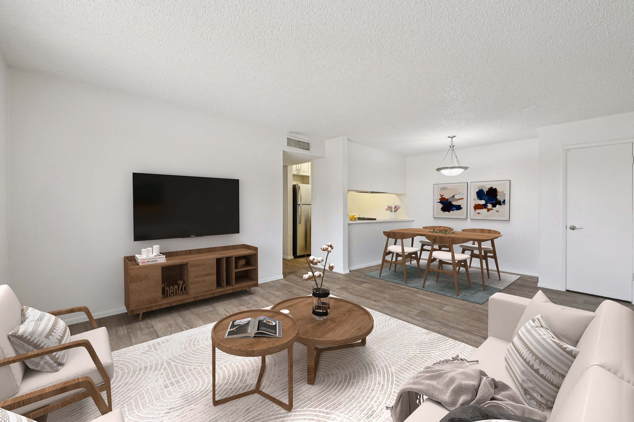 A modern living room featuring a television against a white wall, a wooden TV stand, and two beige armchairs. A coffee table with magazines sits on a textured rug. In the background, a dining area with a round table and chairs is visible, along with a small open kitchen. The decor includes artwork and plants.