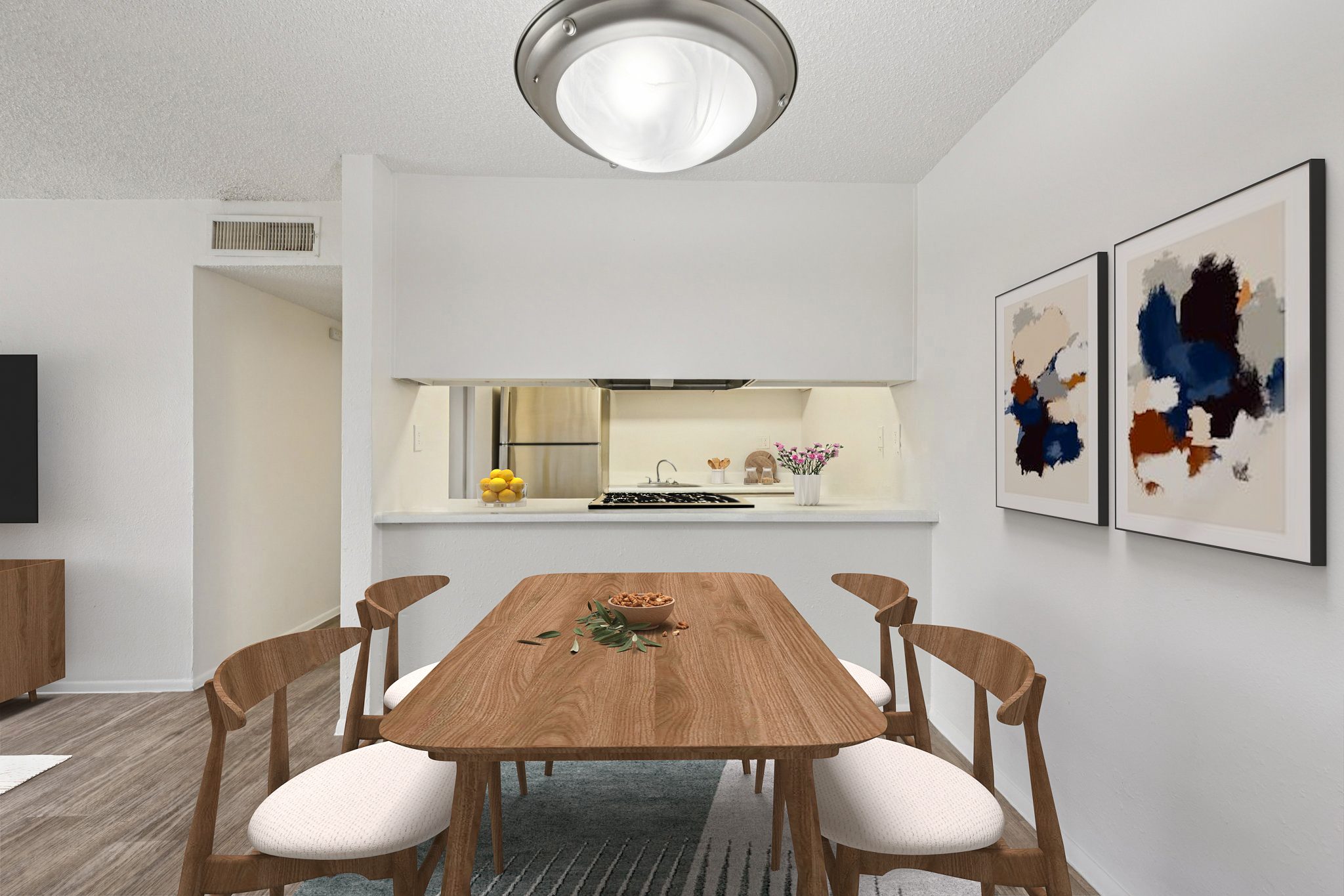 A modern dining area featuring a wooden table with light-colored chairs, elegantly set with a bowl of nuts. The room has a minimalist aesthetic with white walls, a light fixture overhead, and two abstract framed art pieces on the wall. A small kitchen area with a countertop is visible in the background.