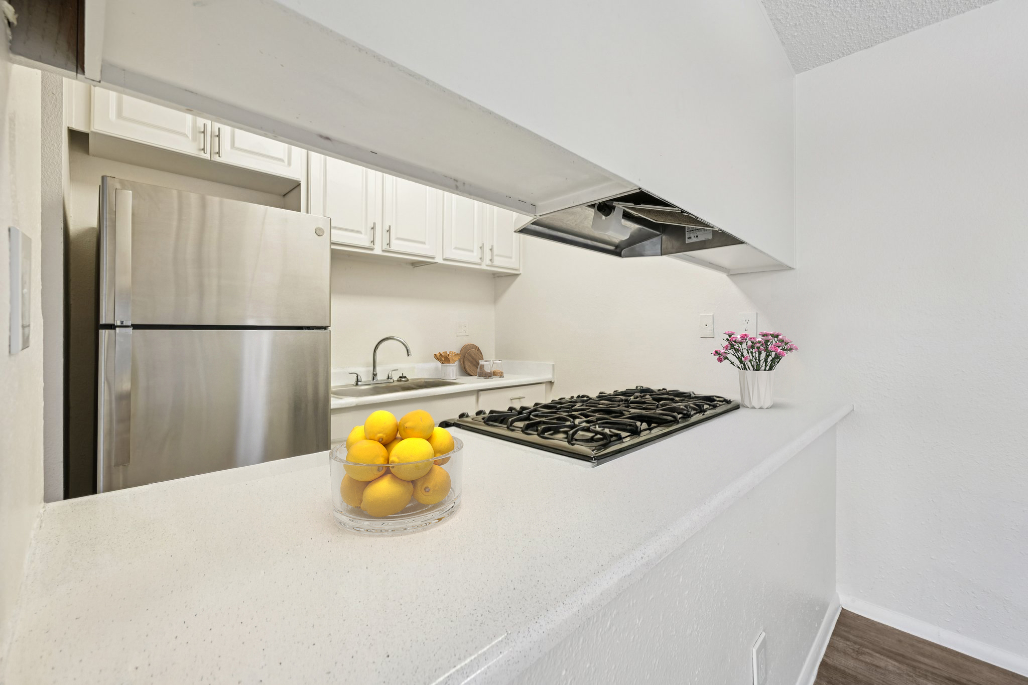 Modern kitchen with a white countertop and a built-in gas stove. A glass bowl filled with yellow lemons sits in the foreground. Stainless steel refrigerator and white cabinets are visible in the background, creating a bright and clean aesthetic. A small vase with flowers adds a touch of color.