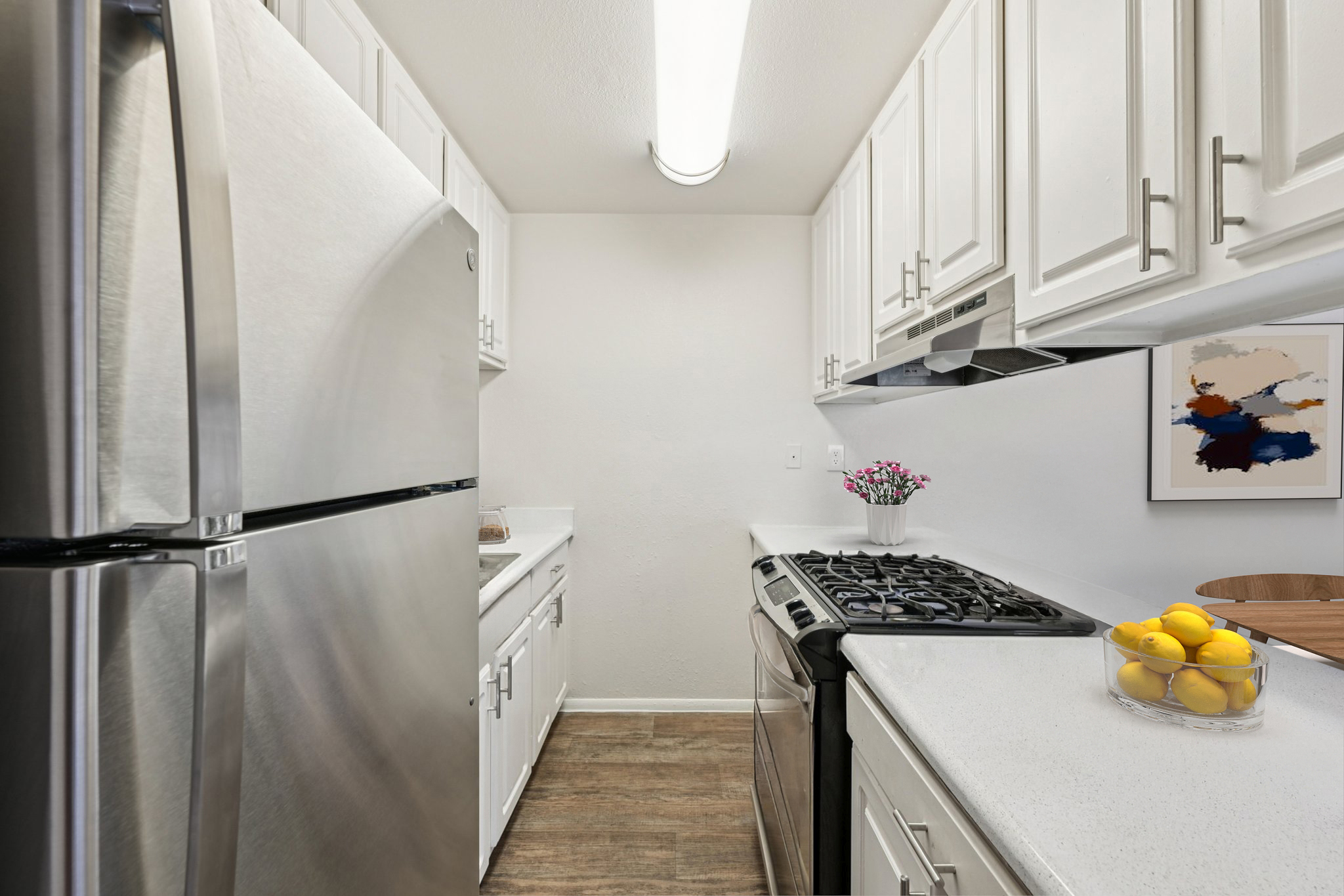 A modern kitchen featuring stainless steel appliances, white cabinetry, and a light countertop. A gas stove is visible, and a bowl of lemons sits on the counter. The kitchen is well-lit with overhead lighting and includes decorative items such as a plant and wall art. The overall aesthetic is clean and bright.