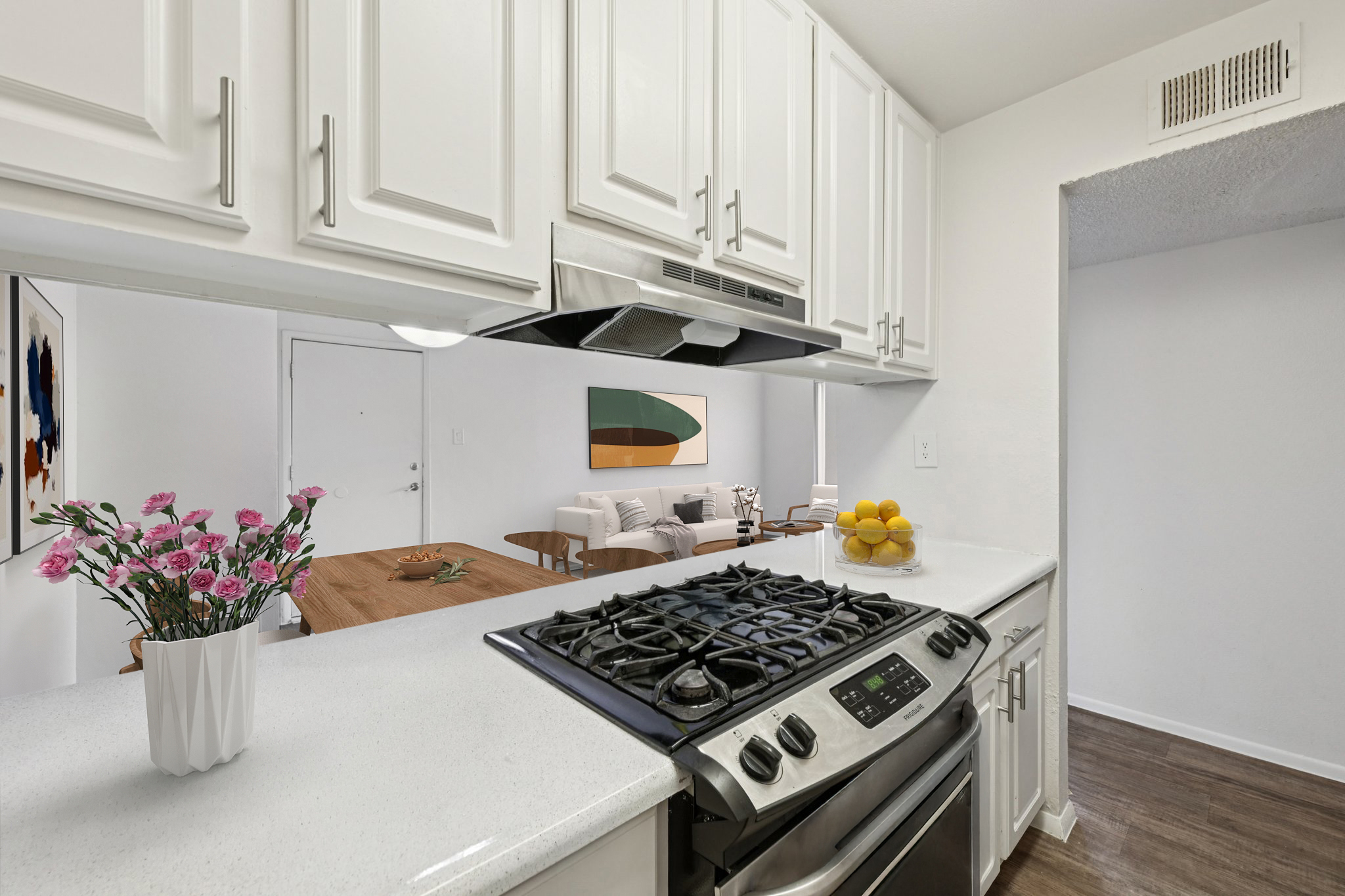 A modern kitchen featuring white cabinets, a stainless steel gas stove, and a white countertop. A vase of pink flowers sits next to a bowl of yellow lemons. In the background, a dining area with a wooden table and minimalist decor is visible. The overall atmosphere is bright and inviting.