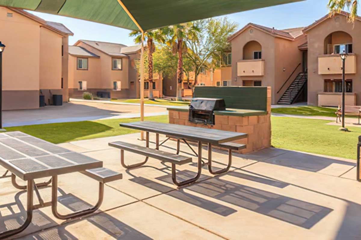 A picnic area with metal tables and a grill under a shaded canopy, surrounded by green grass and palm trees.
