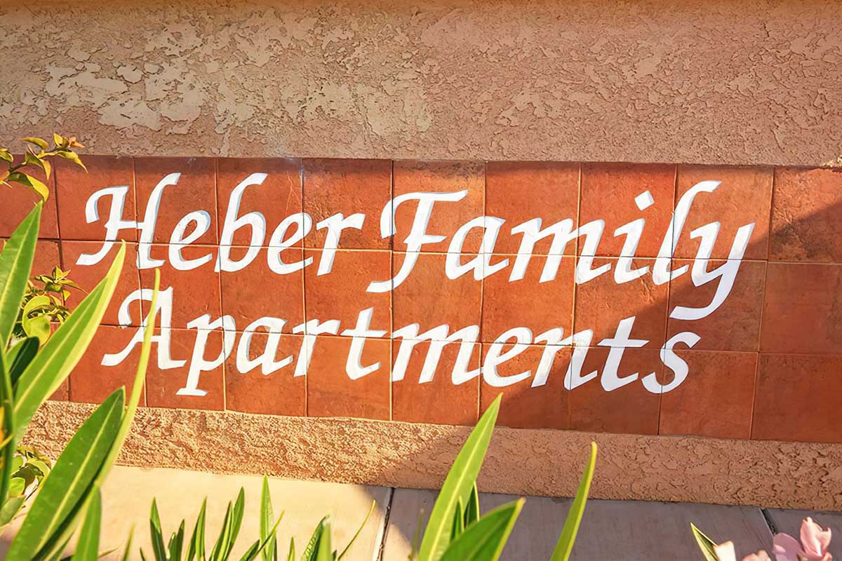 Sign displaying the text "Heber Family Apartments" in white cursive font on a textured brown wall, surrounded by green plants.