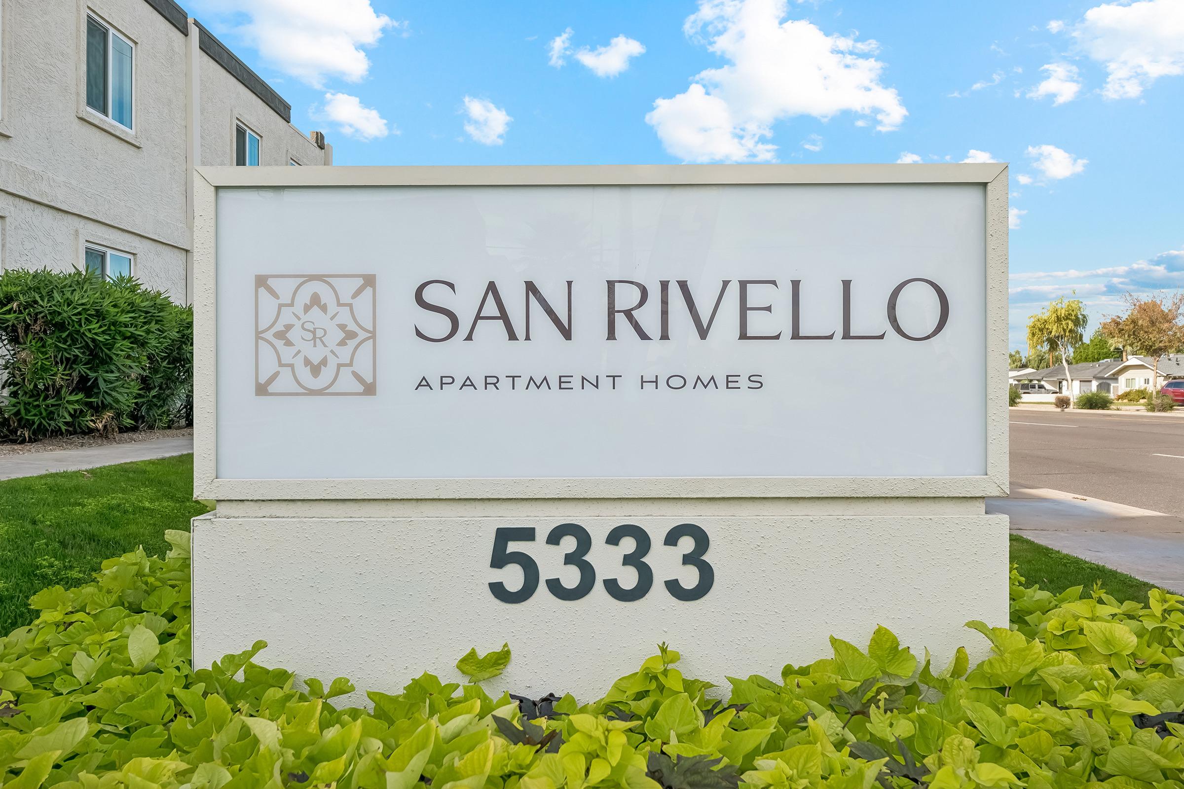 Sign displaying "San Rivello Apartment Homes" with the address "5333." The sign is white with a decorative emblem and surrounded by green plants. The backdrop shows a clear blue sky with fluffy clouds.