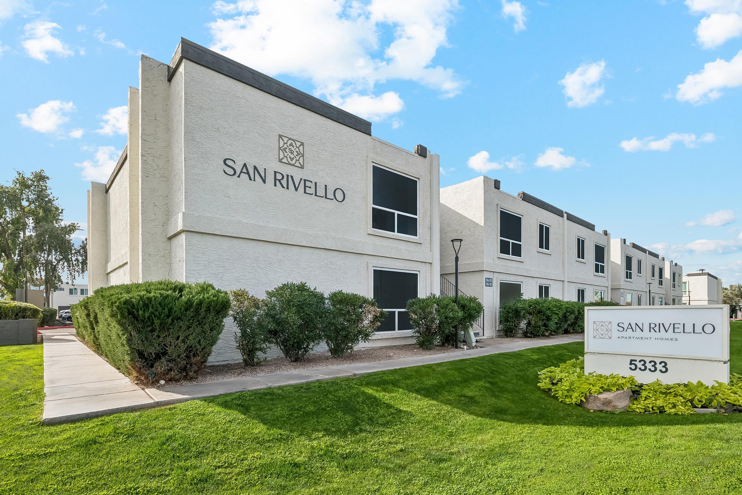Exterior view of San Rivello apartment complex, featuring a modern design in light-colored stucco. The building is surrounded by neatly trimmed shrubs and a manicured lawn. A white sign displays the name "SAN RIVELLO" and the address "5333." The sky is clear with a few scattered clouds.
