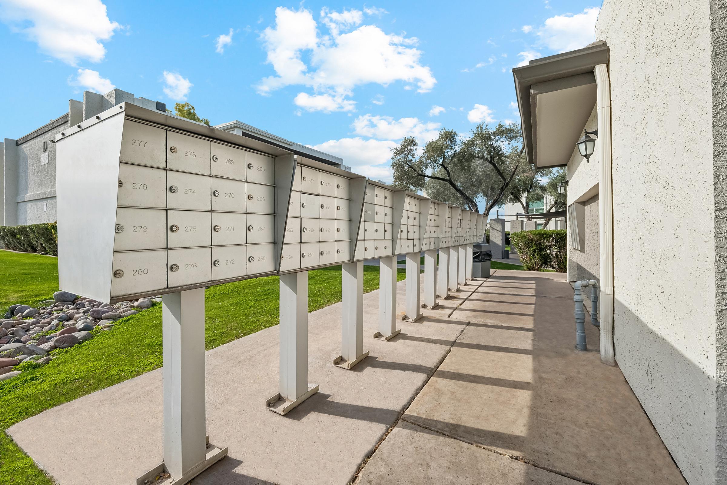 A row of metallic mailboxes on sturdy stands, lined up along a pathway beside a residential building. The area features green grass and trees, with a clear blue sky and fluffy clouds in the background.