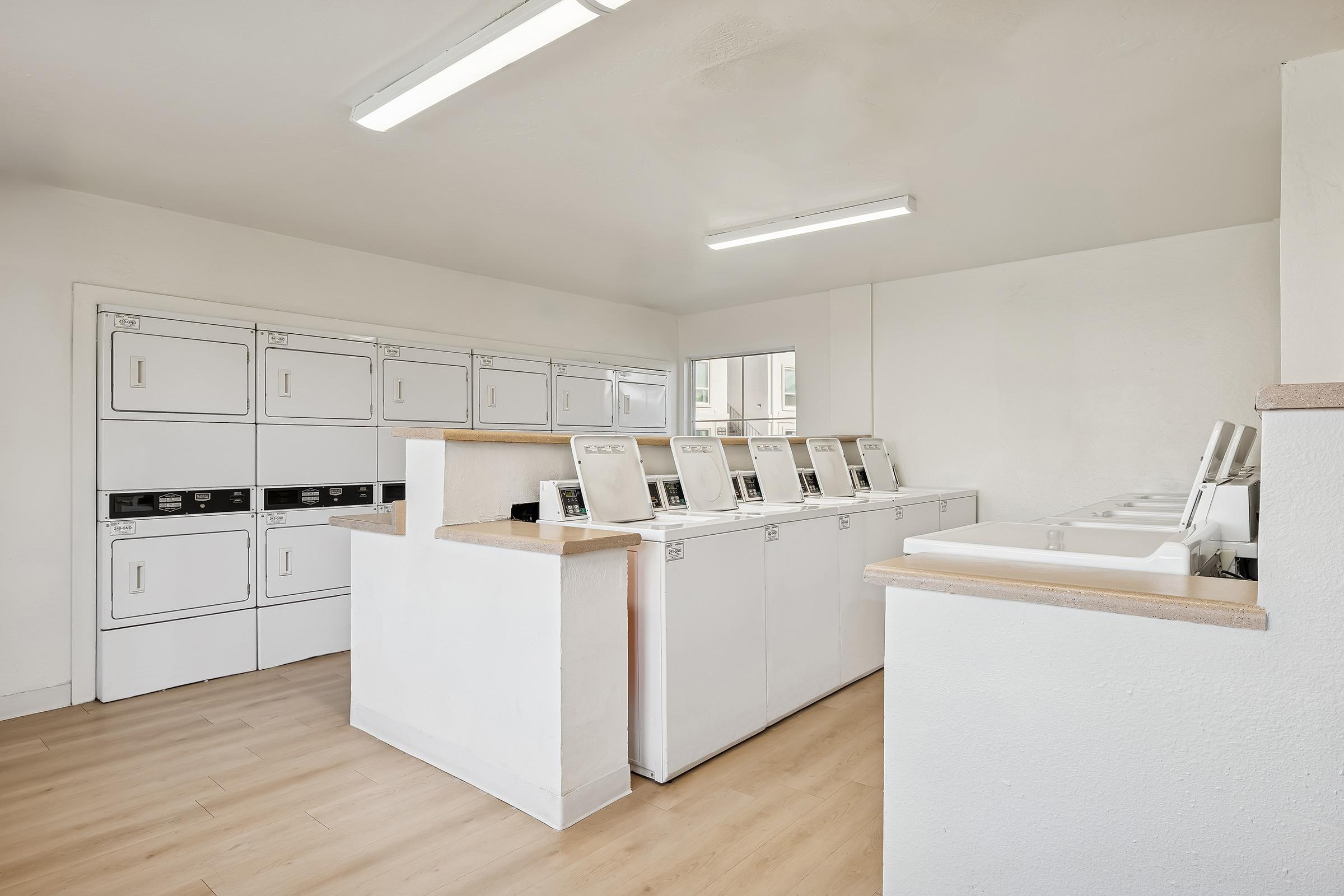 A bright and clean laundry room with multiple stacked washing machines and dryers, arranged in a row against white walls. The space features ample lighting, wood-like flooring, and a service counter. A small window allows natural light to enter, creating an inviting atmosphere for laundry activities.