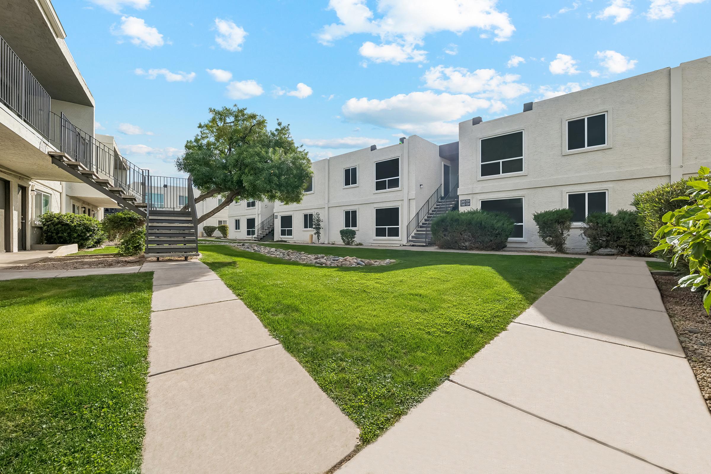A landscaped courtyard with grassy areas and concrete pathways leading between white apartment buildings. There are small shrubs and a tree, with stairs visible on one side. The sky is partly cloudy, adding a bright, inviting atmosphere to the scene.