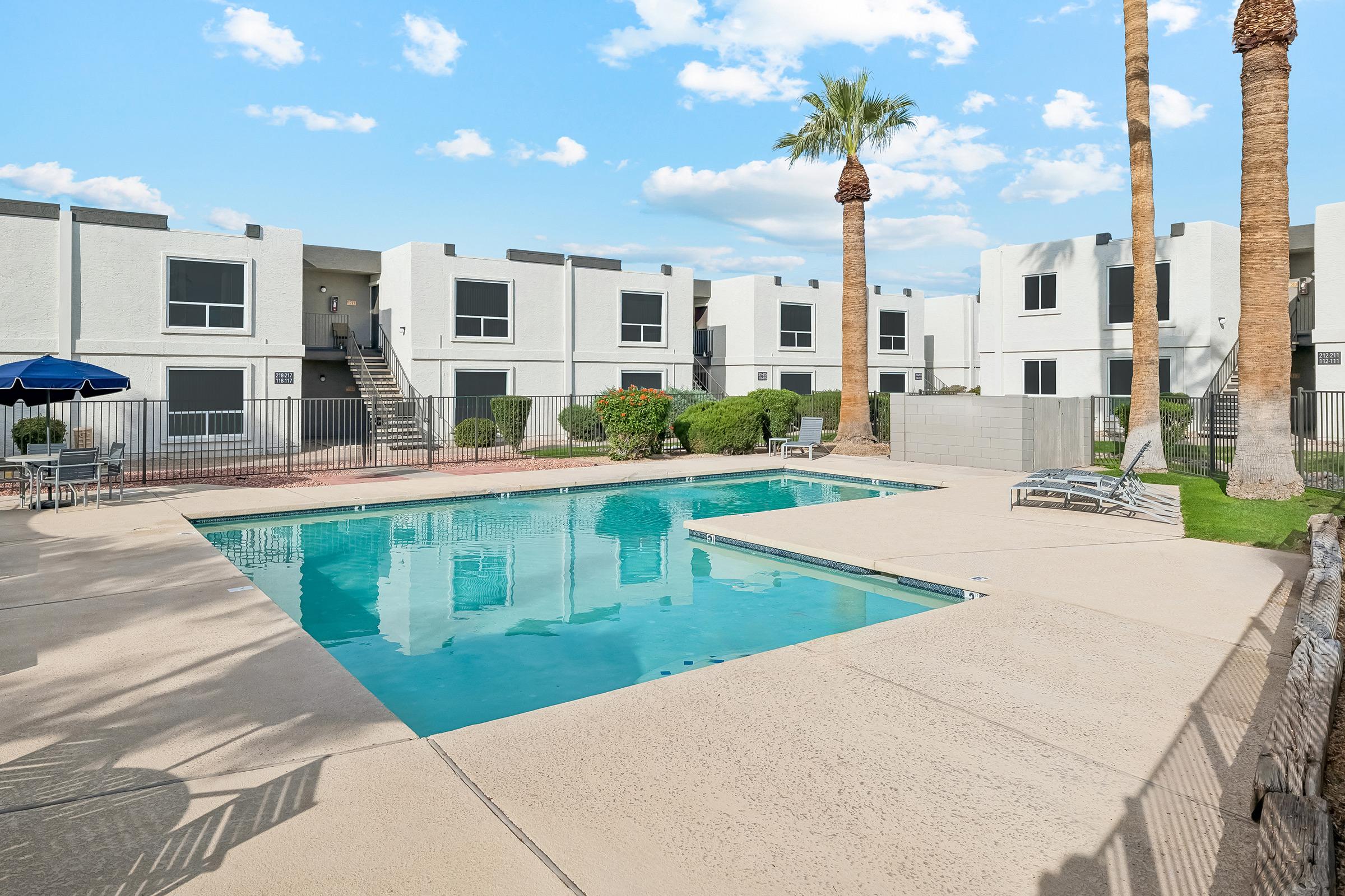 A clear swimming pool surrounded by a spacious outdoor area, featuring lounge chairs and palm trees. In the background, multiple white apartment buildings with balconies can be seen. The sky is bright blue with scattered clouds, creating a sunny atmosphere.