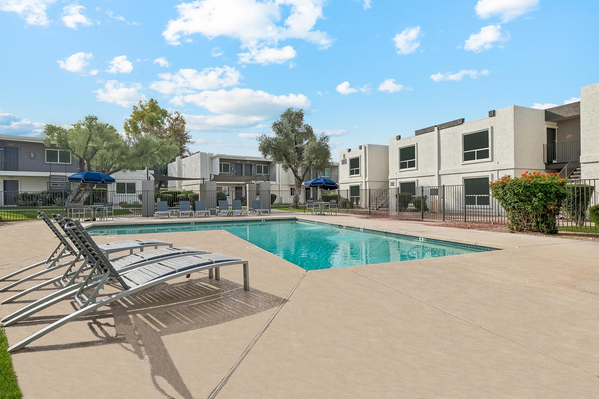 A sunny residential pool area featuring a clear blue pool surrounded by lounge chairs, with shaded seating options under umbrellas. In the background, there are modern apartment buildings and greenery, creating a relaxing outdoor environment. Fluffy white clouds are visible in a bright blue sky.