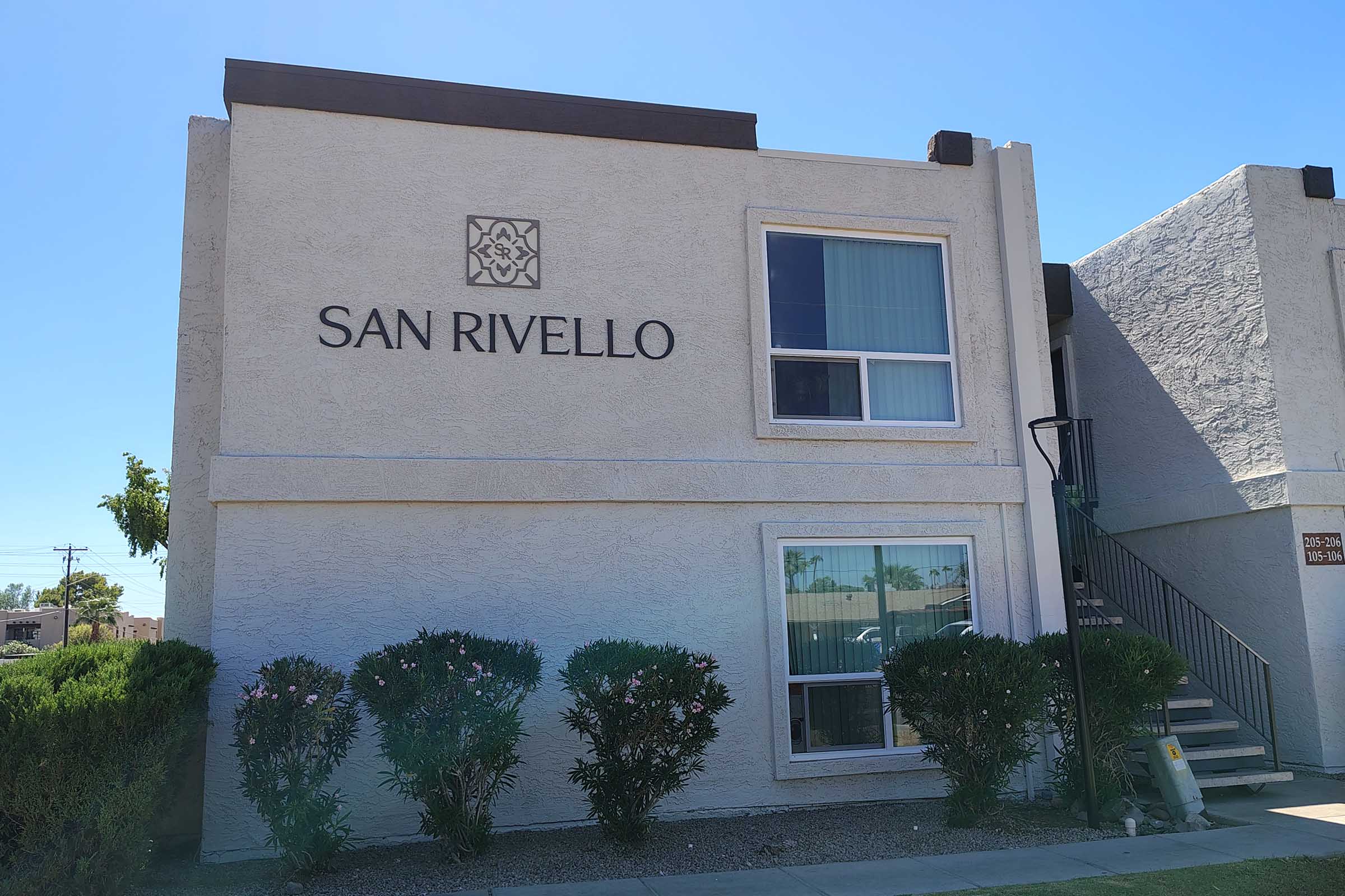 A exterior view of a multi-story residential building with the name "SAN RIVELLO" displayed prominently on the upper wall. The building features large windows, a staircase on the side, and landscaped shrubs in front. The sky is clear and sunny.