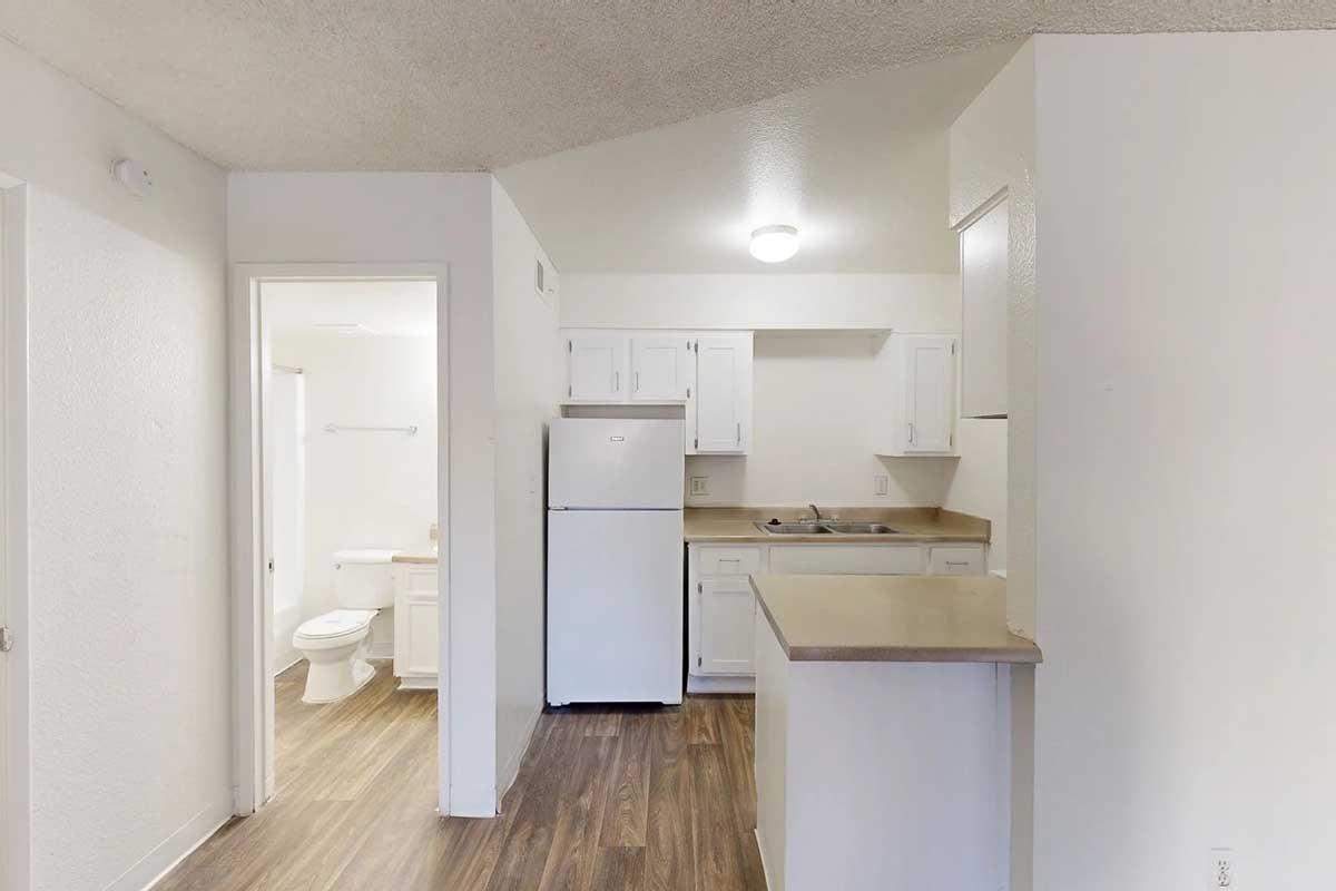 View of a small kitchen area in an apartment, featuring white cabinets, a refrigerator, sink, and countertop. In the background, a partially visible bathroom with a toilet and sink can be seen through an open door. The flooring is a wooden laminate, and the ceiling has a light fixture.
