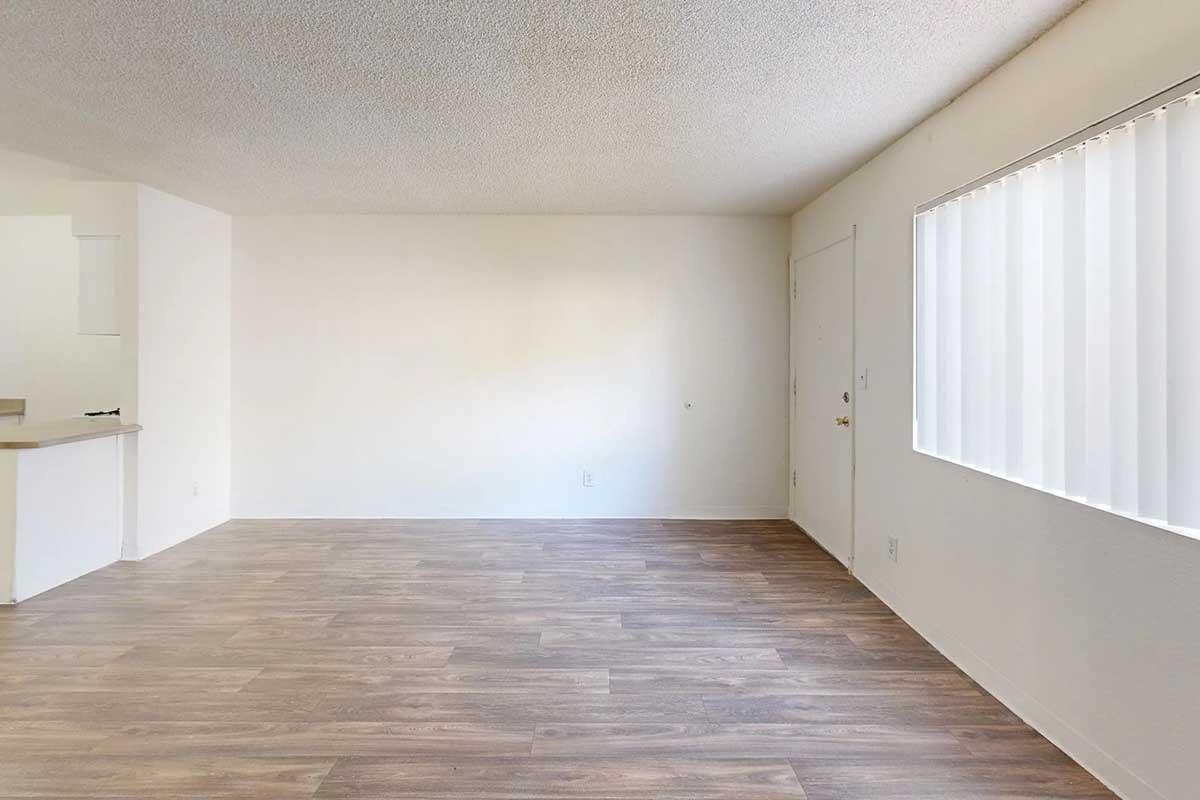 Empty room with light-colored walls and wooden laminate flooring. A door is visible on one side, and there is a large window with vertical blinds letting in natural light. The room has a minimalist appearance with no furniture or decorations.