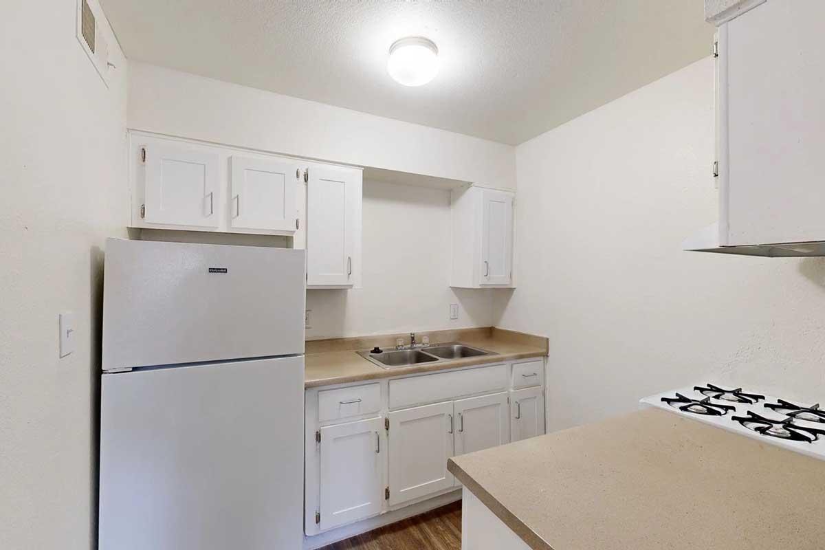A small kitchen featuring white cabinets, a countertop, a stainless steel sink, a refrigerator, and a gas stove. The walls are light-colored, and the floor appears to be a wood laminate. A ceiling light illuminates the space.