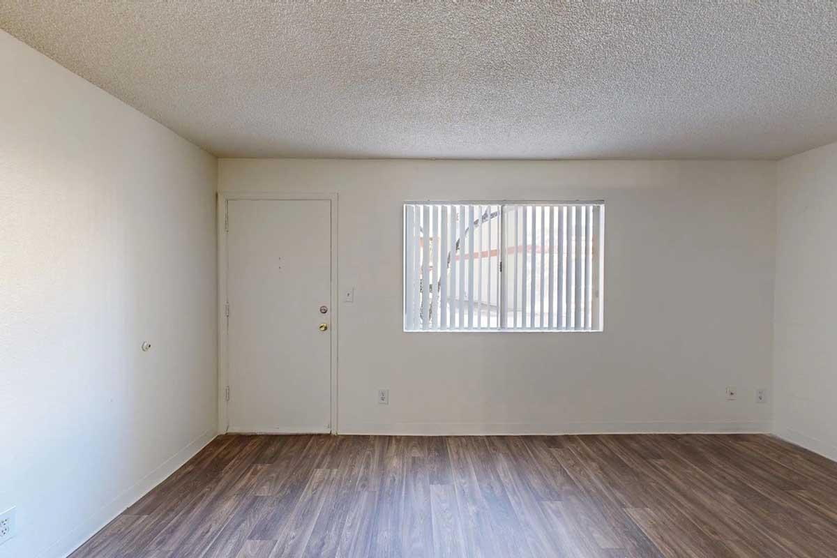 A vacant room with light-colored walls and a textured ceiling. The floor is covered in laminate wood, and there is a single window with vertical blinds allowing natural light. A door is visible on the left side of the image. The space appears clean and unoccupied, ready for decoration or furniture arrangement.