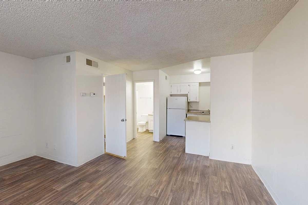 Interior view of a small, unfurnished apartment. The space includes a kitchen area with a refrigerator, a doorway leading to a bathroom, and a living area with laminate flooring. The walls are painted white, and the ceiling has a textured finish. Natural light enters from an unseen window.