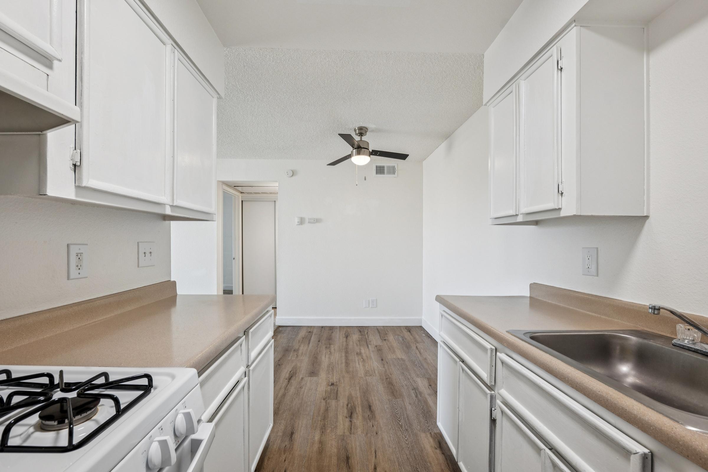 Interior view of a kitchen featuring white cabinets, a beige countertop, and a stainless steel sink. A gas stove is visible on the left, and the kitchen has a spacious, open layout leading to a well-lit living area with a ceiling fan. The floor is made of wood laminate.