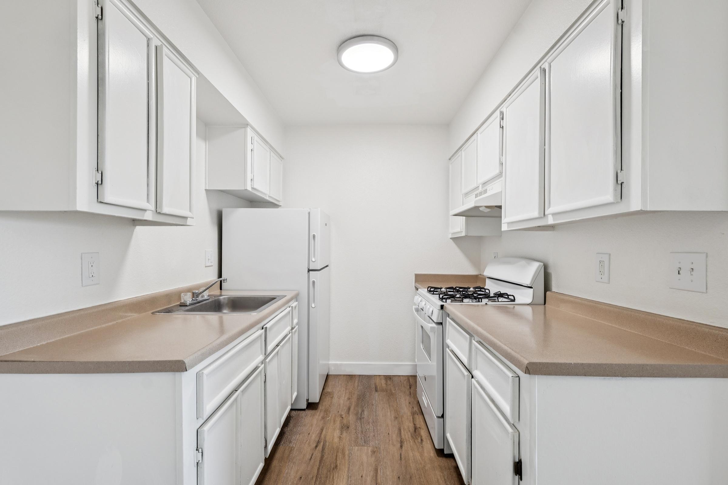 A modern kitchen featuring white cabinetry, a stainless steel sink, a refrigerator, and a gas stove on a light-toned countertop. The walls are painted in a neutral color, and there is a flush ceiling light for illumination. The floor is covered with wooden laminate, creating a clean and bright atmosphere.
