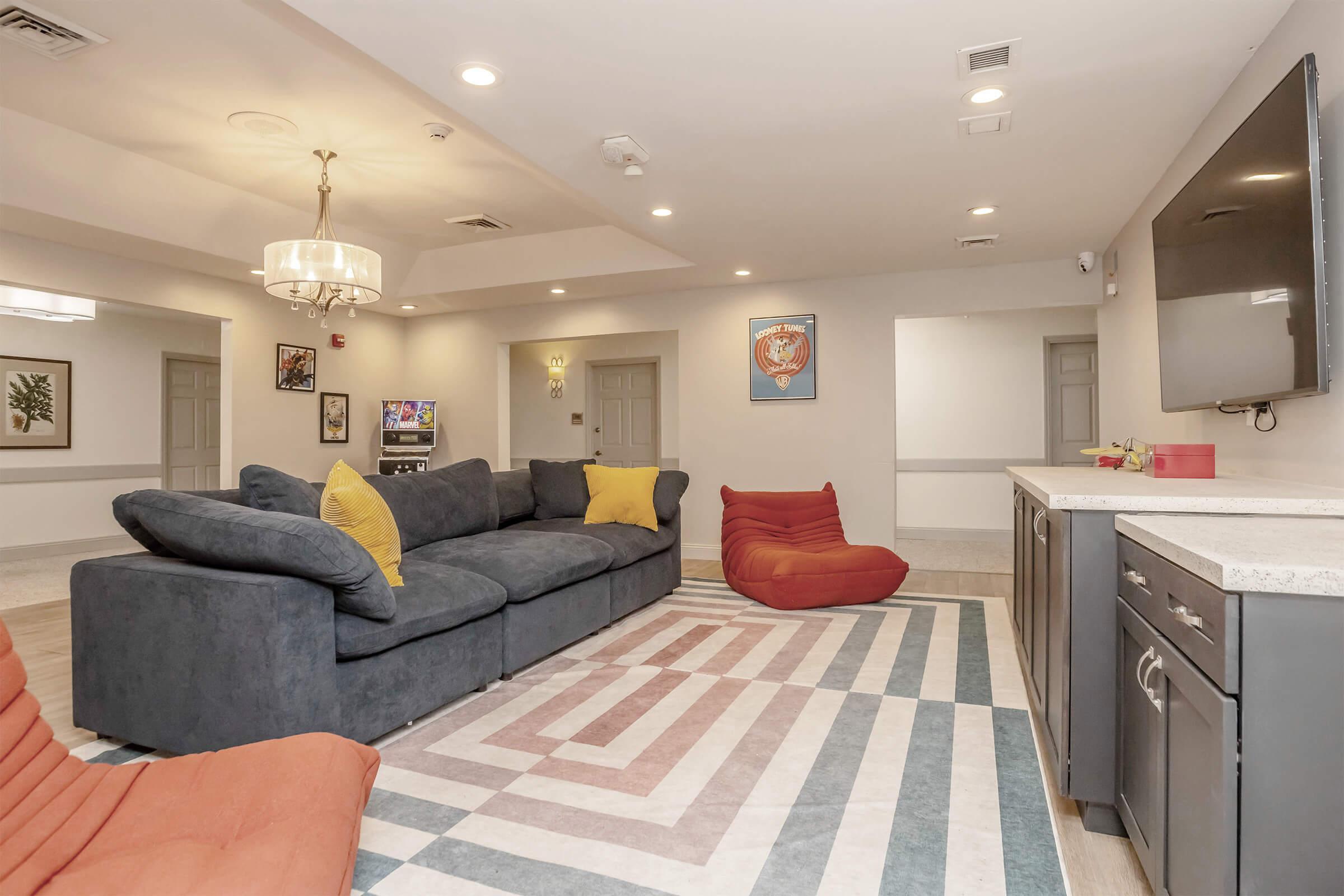 Cozy lounge area featuring a large blue sectional sofa with yellow and gray pillows, a red beanbag chair, a striped area rug, and a flat-screen TV mounted on the wall. The space is bright with modern decor, including framed artwork and overhead lighting fixtures.