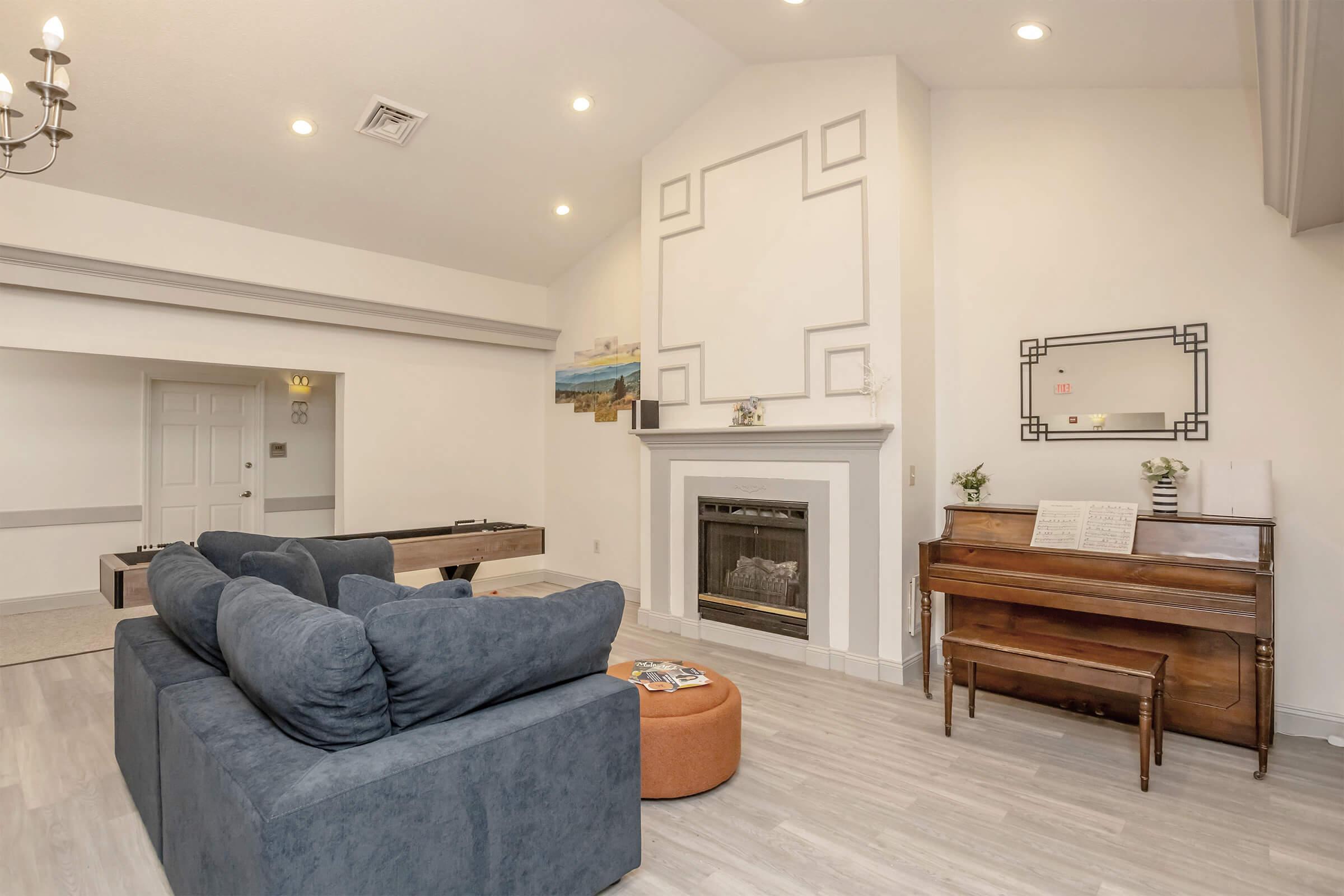 A modern living room featuring a blue sectional sofa, an orange ottoman, and a wooden piano. The walls are painted in neutral tones, with a fireplace and a decorative mirror. Bright overhead lights illuminate the space, enhancing the inviting atmosphere. A small table and a door to another room are also visible.