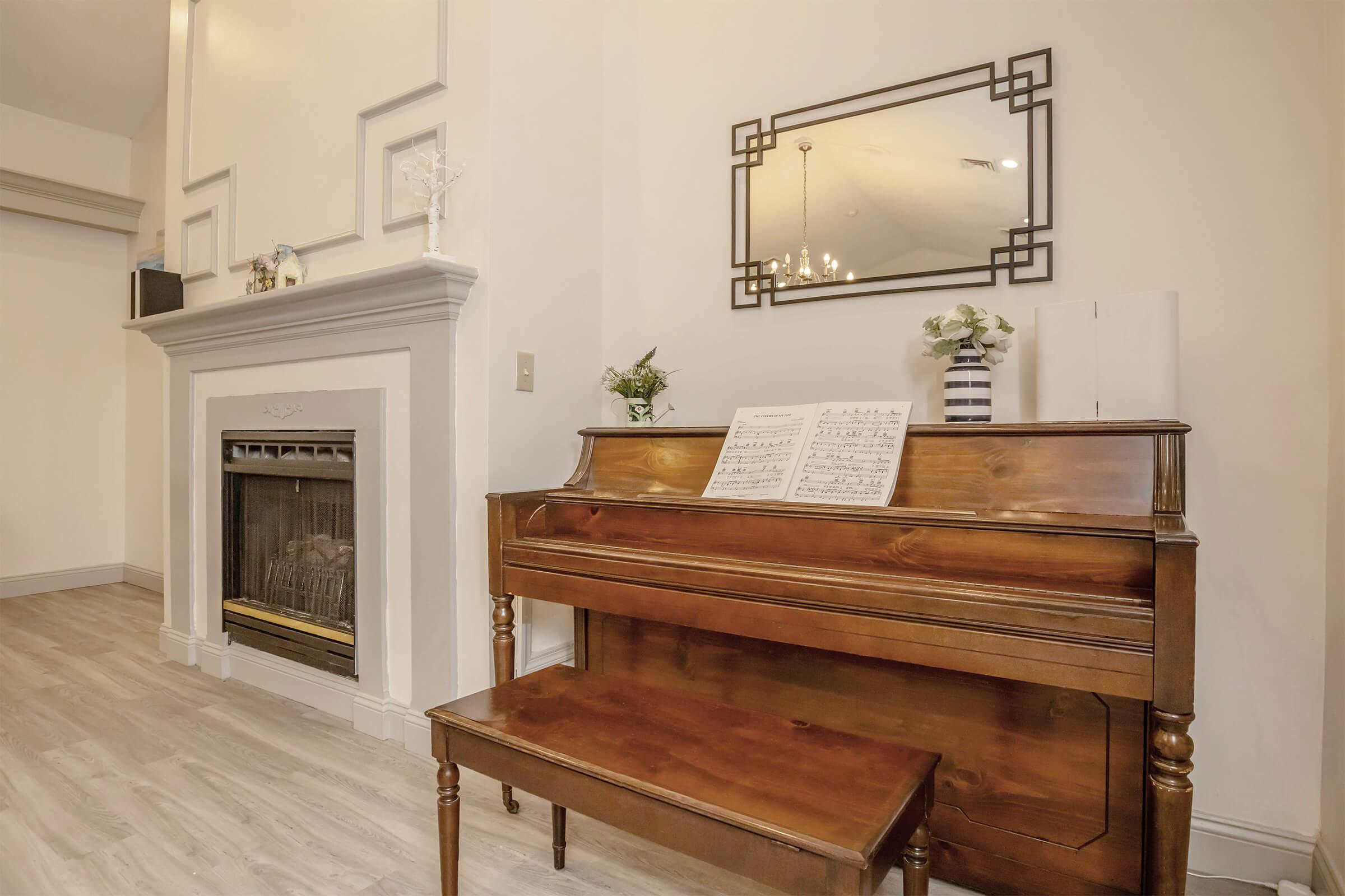 A wooden piano with a matching bench is positioned beside a fireplace in a well-lit room. Sheet music is displayed on the piano. The room features a decorative mirror and is accentuated by simple floral arrangements in modern vases. Soft lighting enhances the cozy atmosphere.