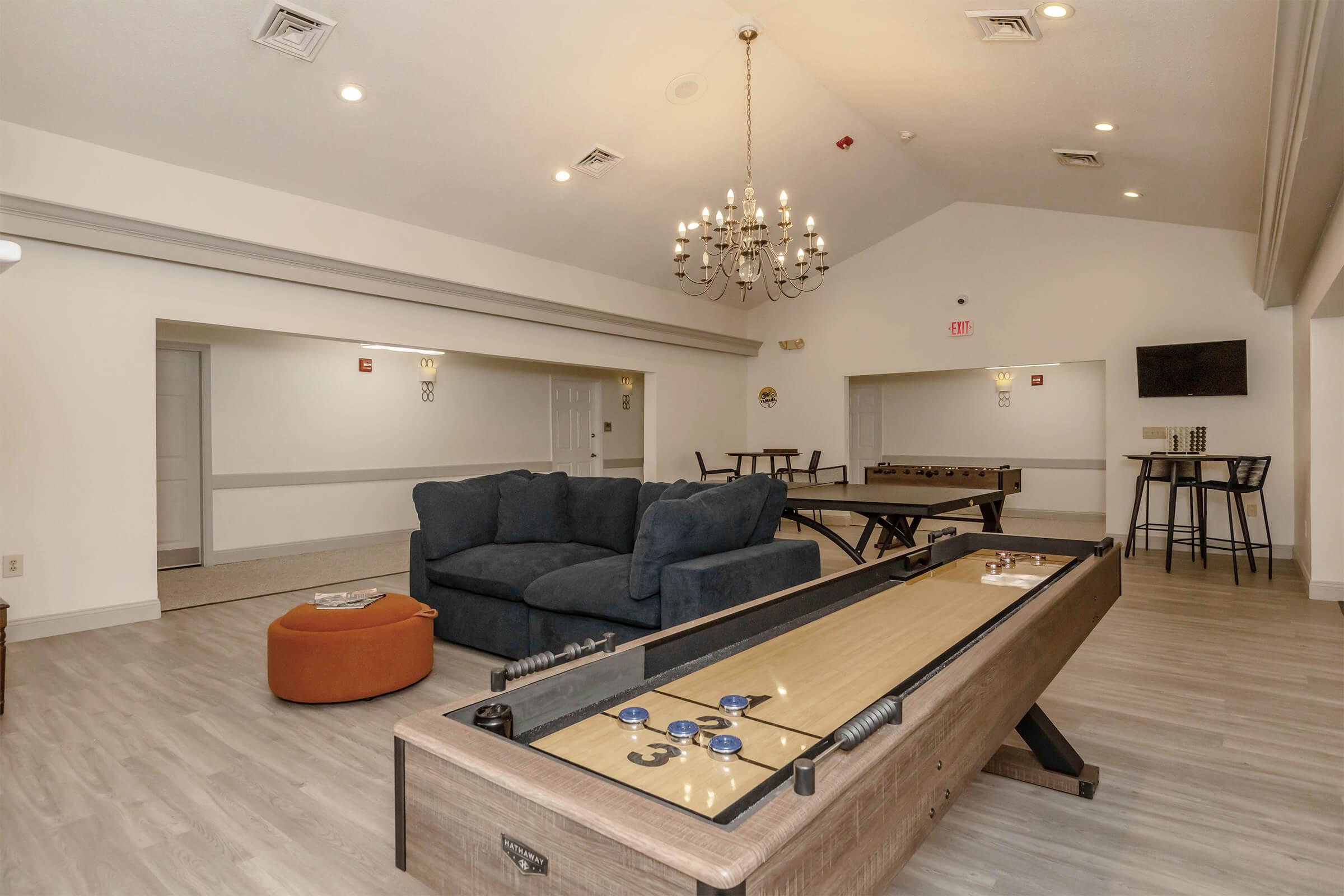 Interior view of a recreational room featuring a shuffleboard table, a dark blue sectional sofa, an orange ottoman, and a chandelier. There are also two game tables in the background and a mounted television on the wall. The space is well-lit and designed for relaxation and entertainment.