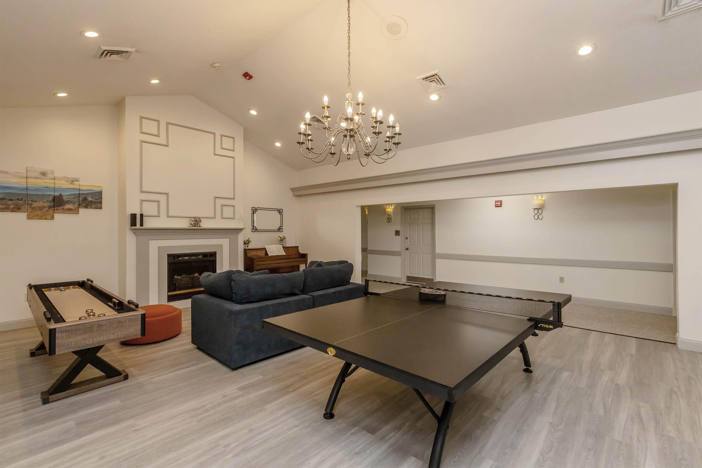 A spacious recreation room featuring a ping pong table, foosball table, and a cozy seating area with a blue couch. A chandelier hangs from the ceiling, and a fireplace is visible in the background. The room has light-colored flooring and neutral walls, creating an inviting atmosphere for leisure activities.