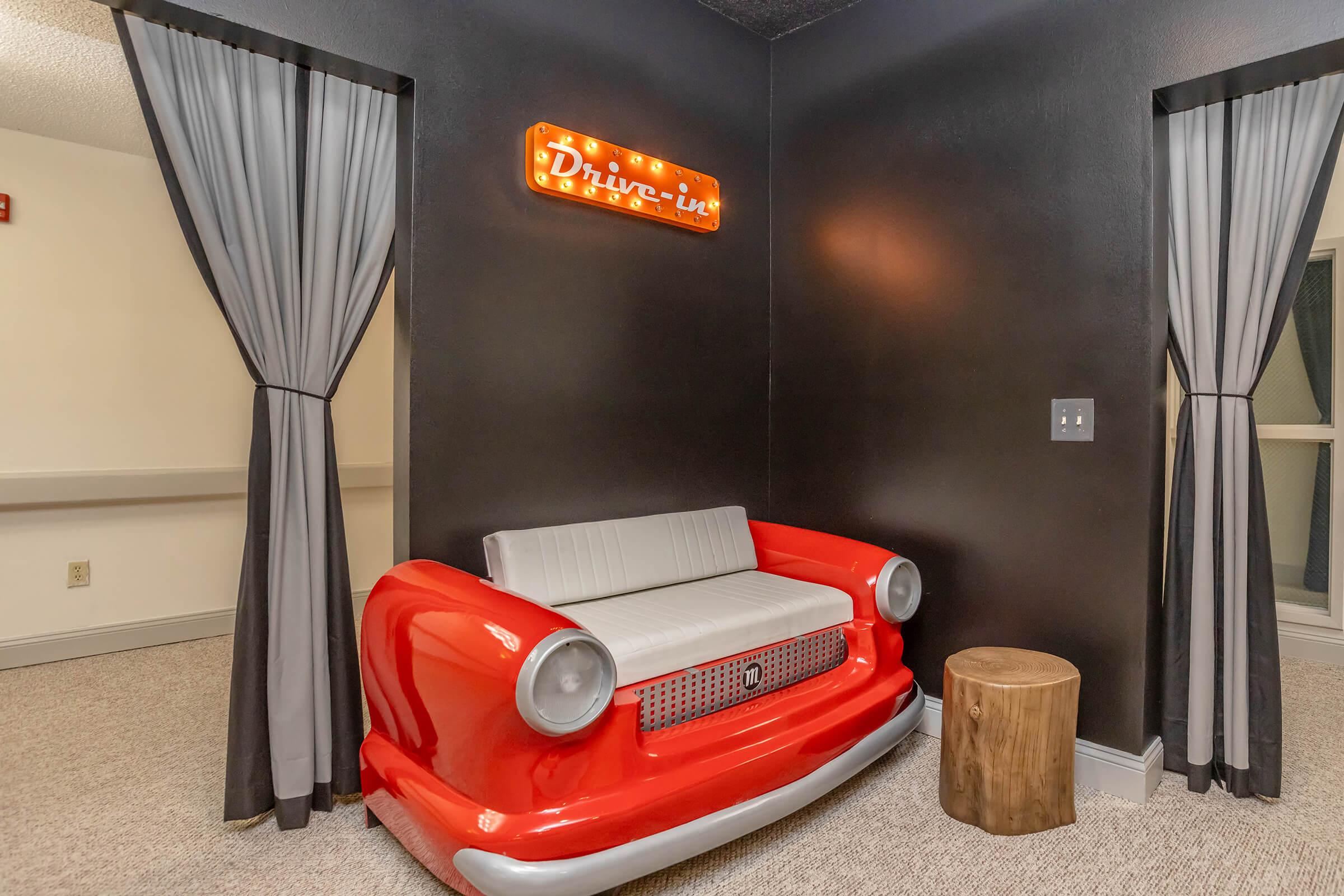 A retro-themed seating area featuring a red car-shaped couch with a gray cushion, set against a dark wall. Above, there’s a vintage-style "Drive-in" sign with lights. Two gray curtains frame the space, and a wooden stump serves as a side table. The overall design evokes a nostalgic diner atmosphere.