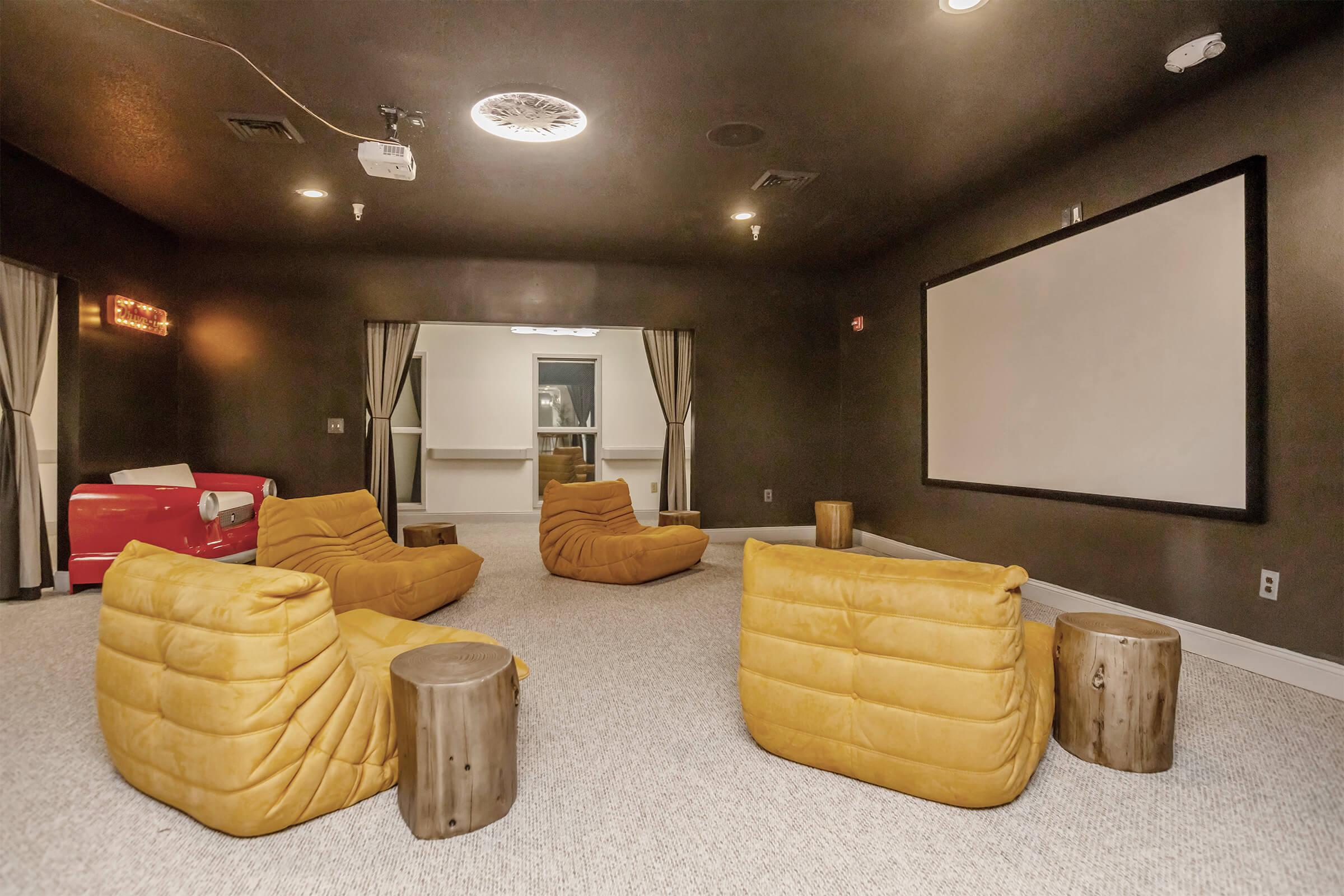 A cozy media room featuring four yellow bean bag chairs arranged around wooden stump tables. A large, blank projection screen is mounted on the wall, and soft lighting illuminates the room. Dark walls create an intimate atmosphere, perfect for watching movies or relaxing with friends.