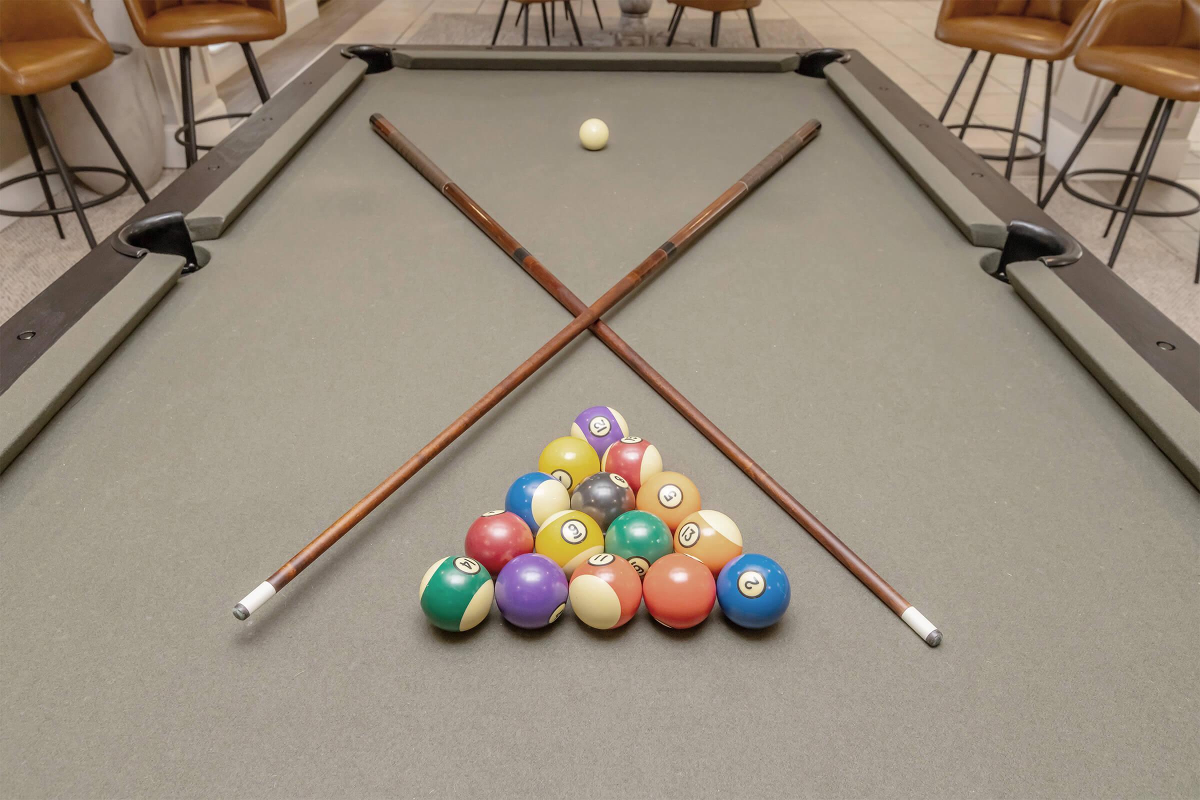 A pool table featuring a triangle of colorful billiard balls arranged for a game, with two cue sticks crossed over the top. A single white cue ball is positioned on the table. The setting includes modern bar stools in the background.