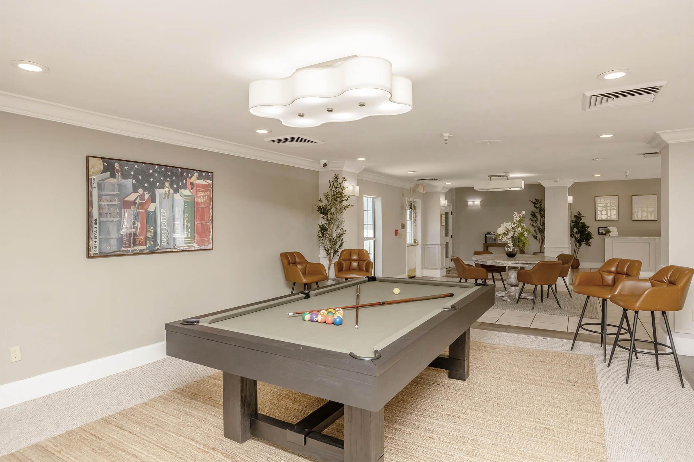 Interior view of a recreational area featuring a pool table with colorful balls on it. There are stylish brown chairs arranged around the room, a decorative rug on the floor, and a wall with framed artwork. The lighting is modern with a distinctive ceiling fixture.