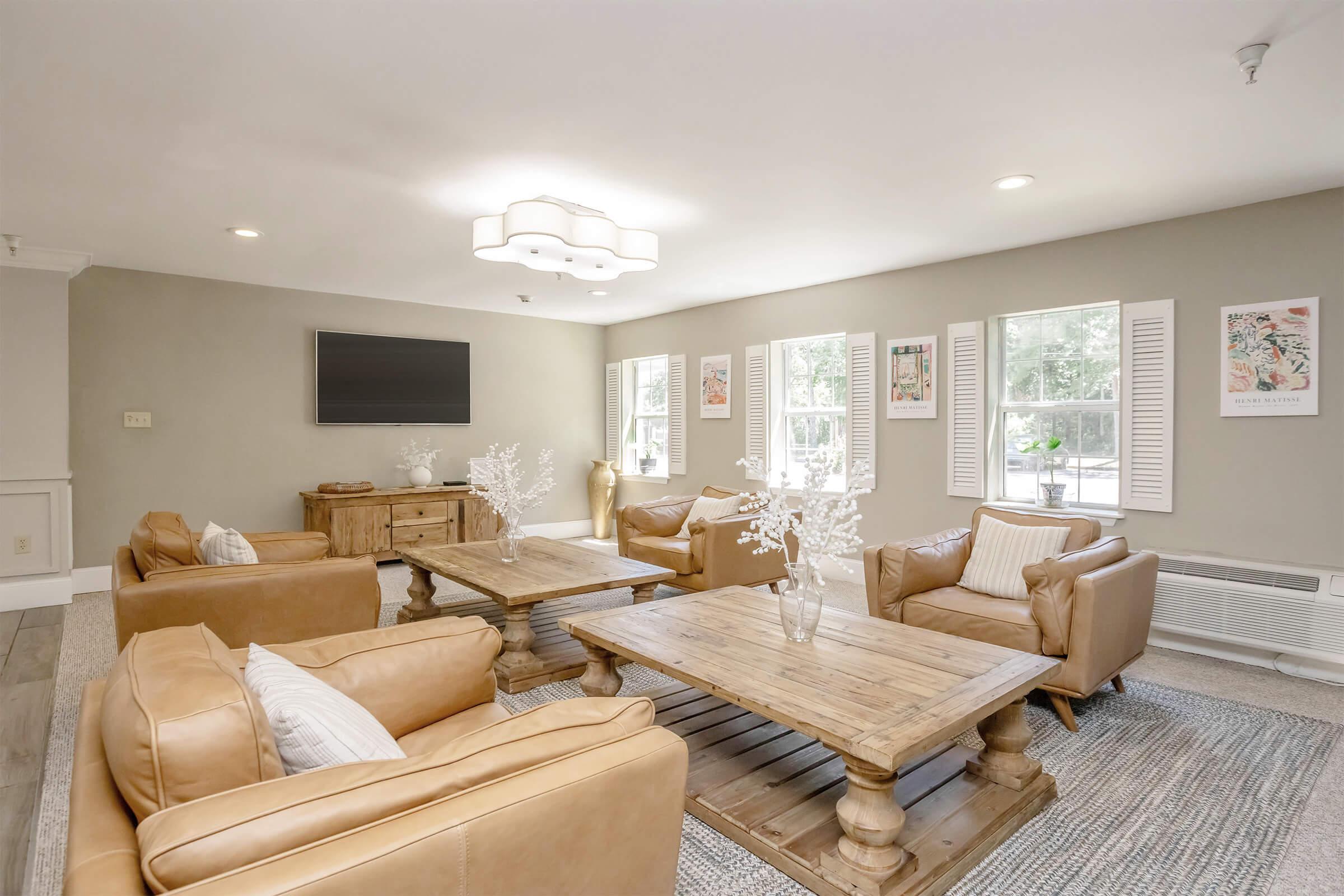 A cozy living room featuring four tan leather armchairs and two wooden coffee tables on a textured rug. There is a flat-screen TV mounted on the wall, and large windows allow natural light to fill the space. The walls are painted a soft gray, with framed artwork and decorative plants adding charm.