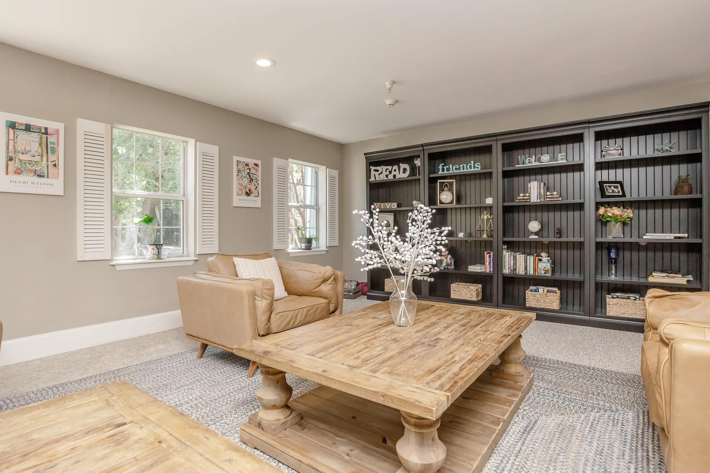 A cozy living room featuring two tan leather chairs, a wooden coffee table, and a large black bookshelf filled with books and decorative items. The walls are painted a soft gray, and there are framed artworks and windows with white shutters. A light rug adds warmth to the space.