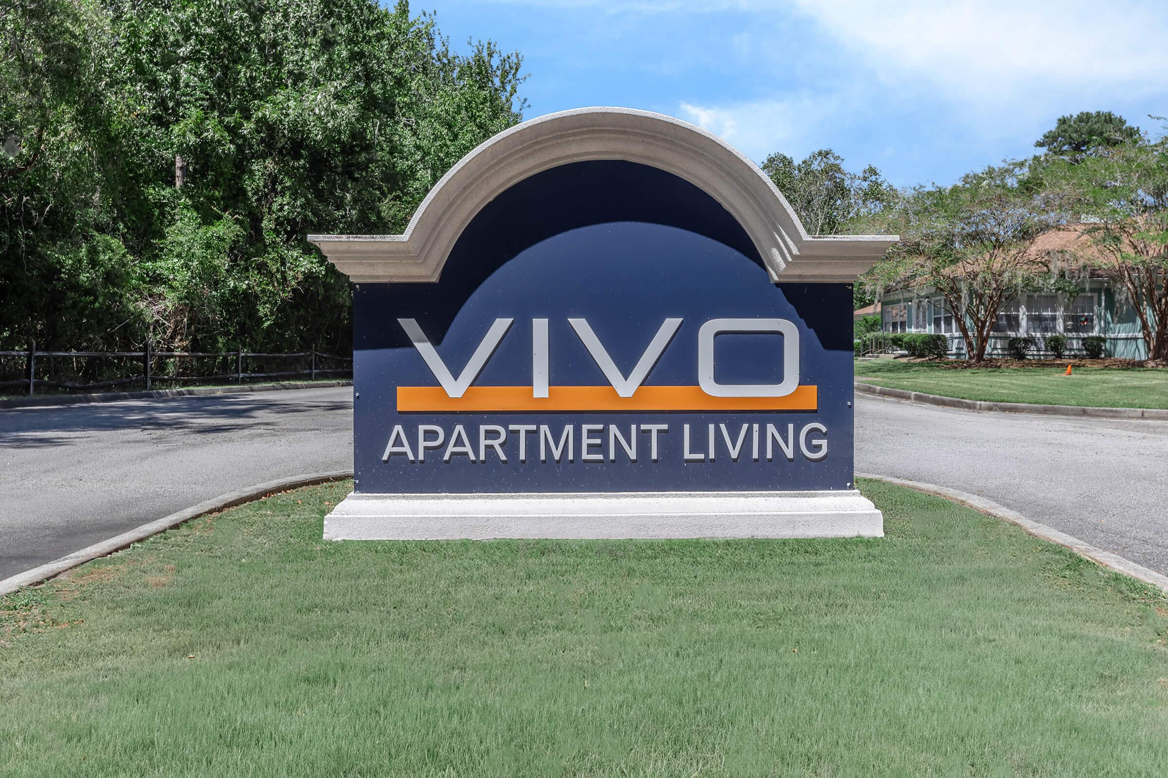 Sign for Vivo Apartment Living, featuring a prominent blue background with white and orange text. The sign is situated at an entrance to the apartment complex, surrounded by green grass and trees under a clear sky.