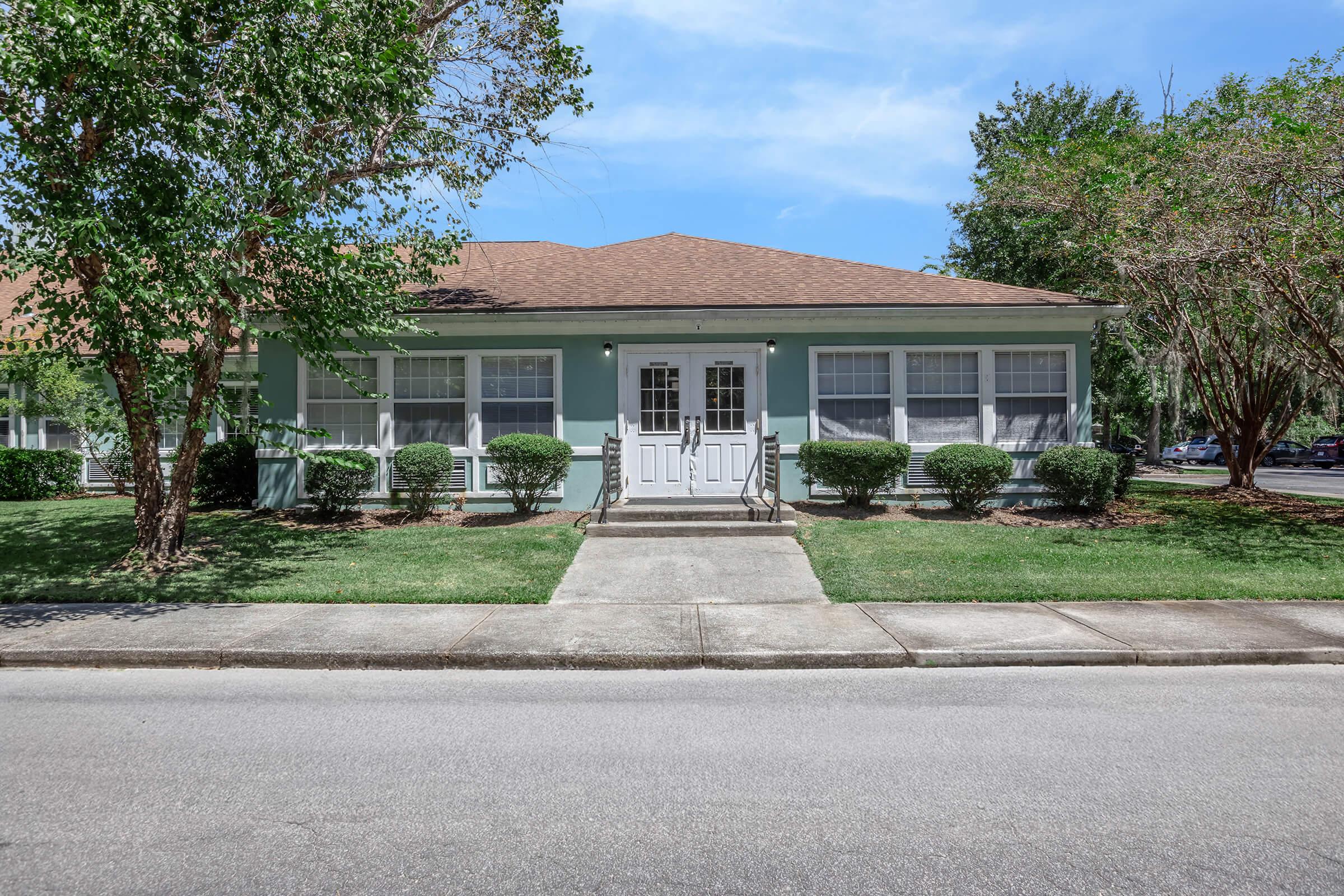 Single-story building with a light blue exterior and a brown roof. It features a symmetrical facade with a central double door and large windows on either side. Neatly trimmed hedges line the front, and a concrete walkway leads to the entrance. Lush green grass and trees are visible in the surroundings.